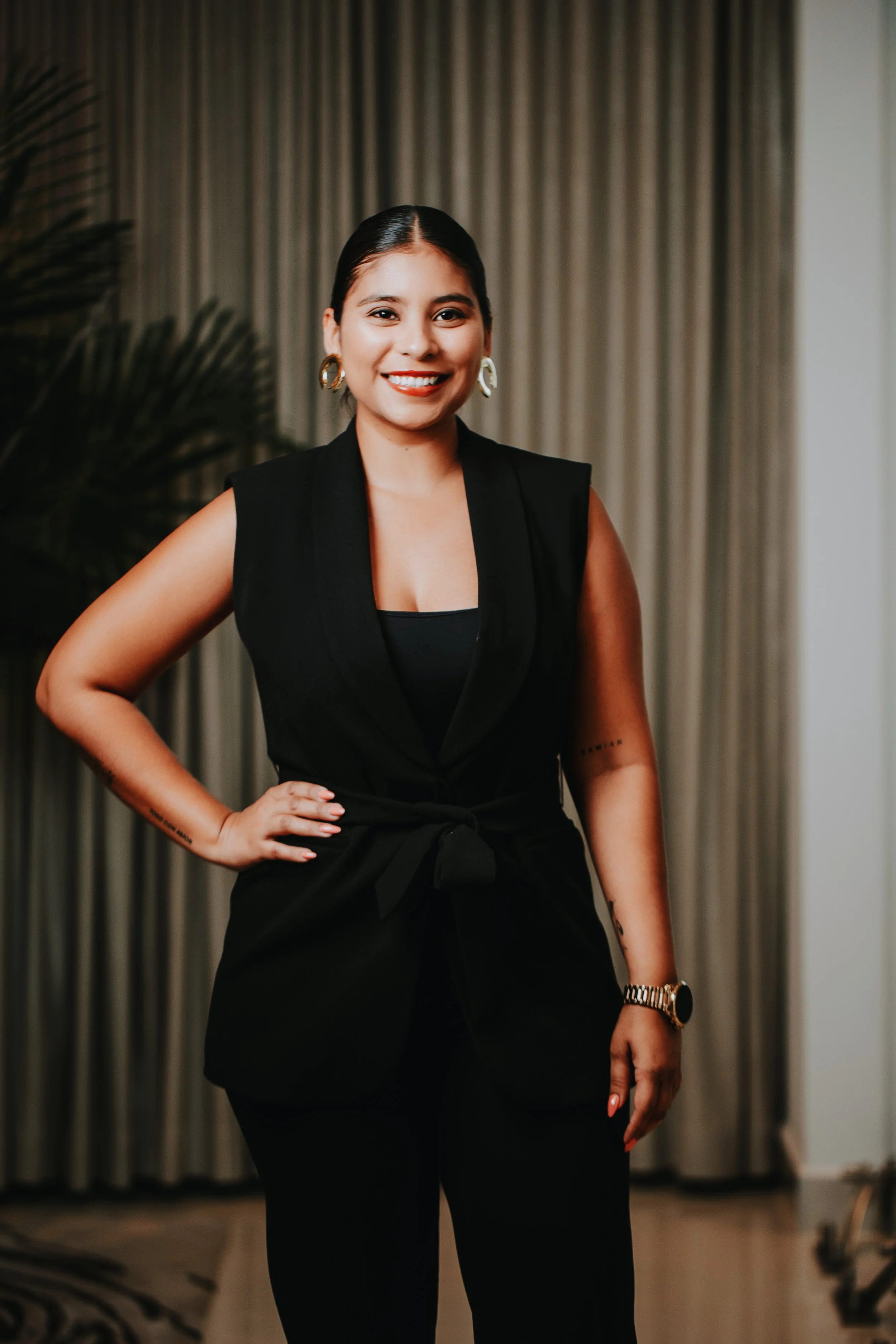 A woman wearing a black sleeveless suit with a waist tie, gold hoop earrings, and a gold wristwatch, posing indoors against a curtain backdrop.