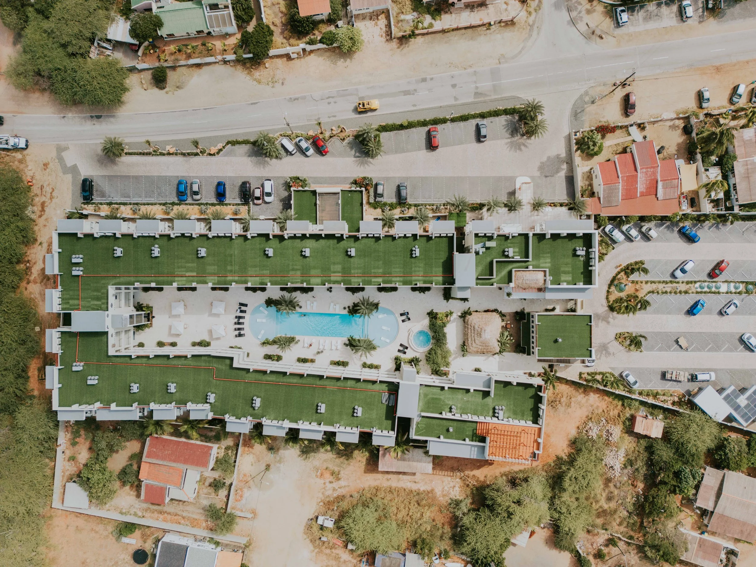 An aerial view of a residential complex with a central swimming pool, surrounded by buildings with green roofs, parking lots, and neighboring houses with red-tiled roofs.