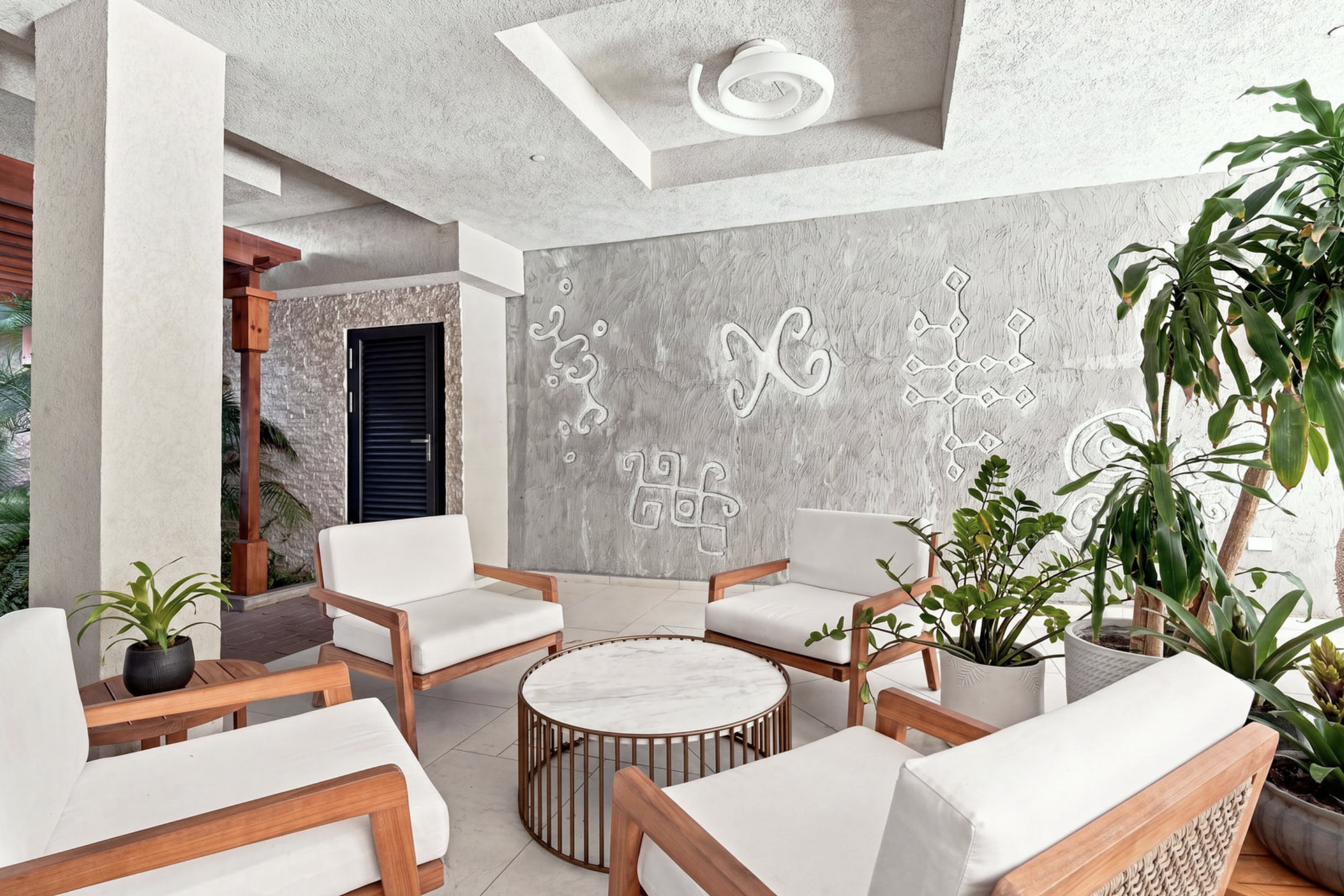 A cozy outdoor seating area with white cushioned chairs and wooden frames surrounded by various green potted plants, a textured gray wall with decorative artwork, and a ceiling light fixture.