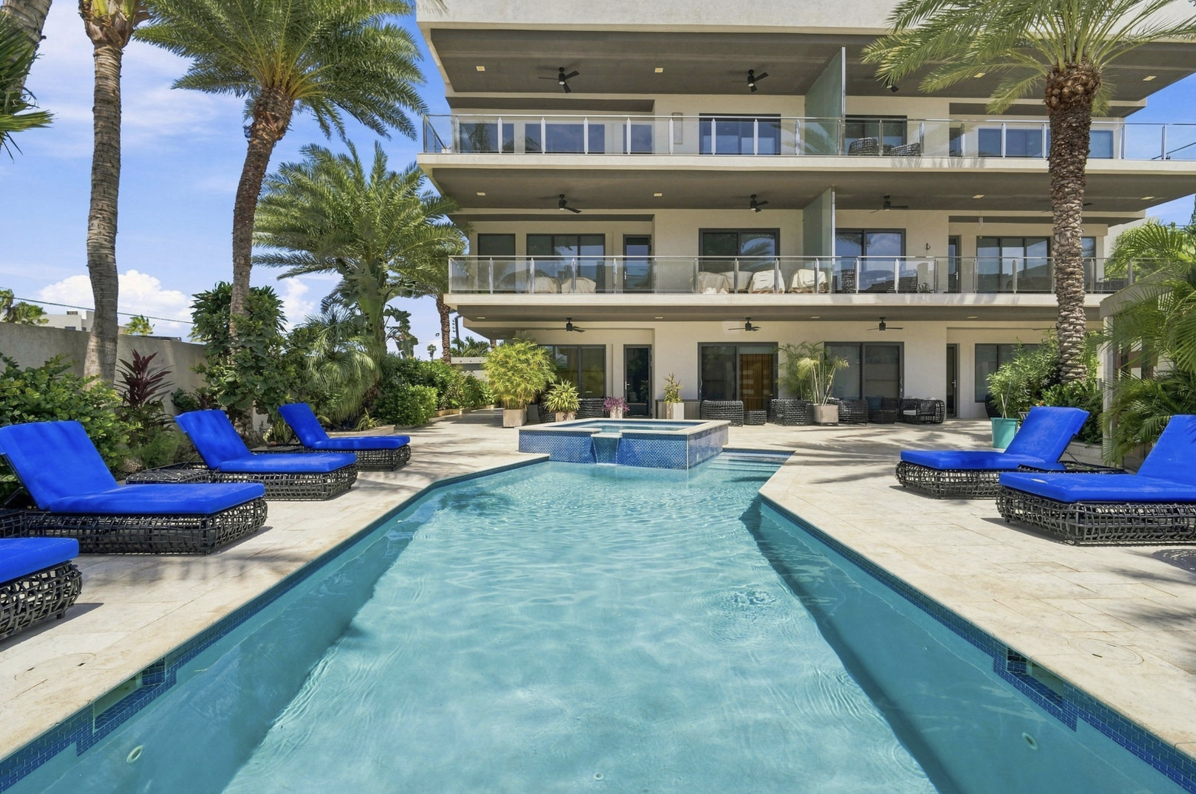 A modern multi-story building with balconies overlooking a backyard pool area surrounded by lush tropical plants and tall palm trees.