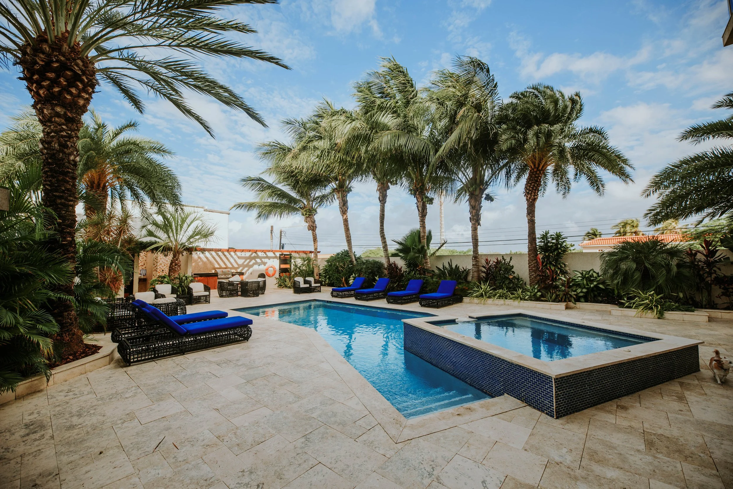 Outdoor pool area with blue lounge chairs, palm trees, and lush greenery under a partly cloudy sky.