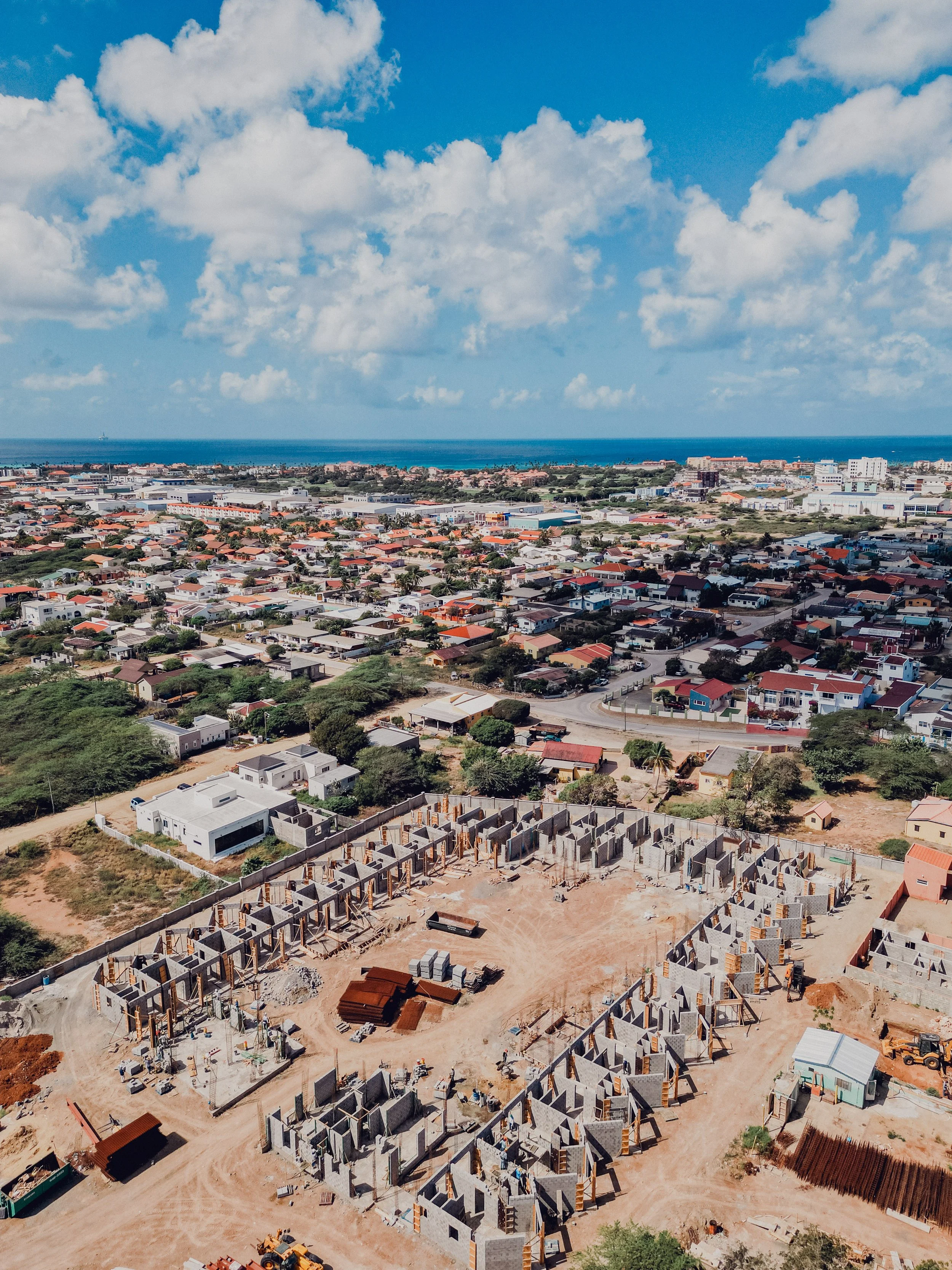 Aerial view of a construction site with partially built houses, surrounded by a residential area with single-family homes, roads, greenery, and the ocean in the background under a partly cloudy sky.
