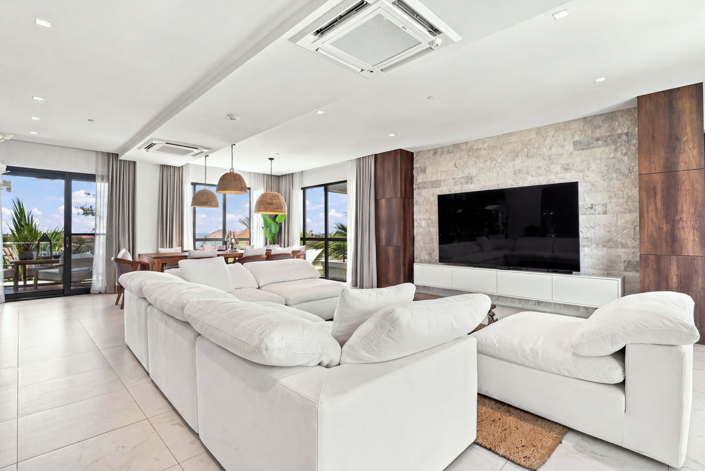 Bright, modern living room with white sectional sofa, large windows with curtains, mounted flat-screen TV, stone accent wall, and hanging wicker light fixtures.