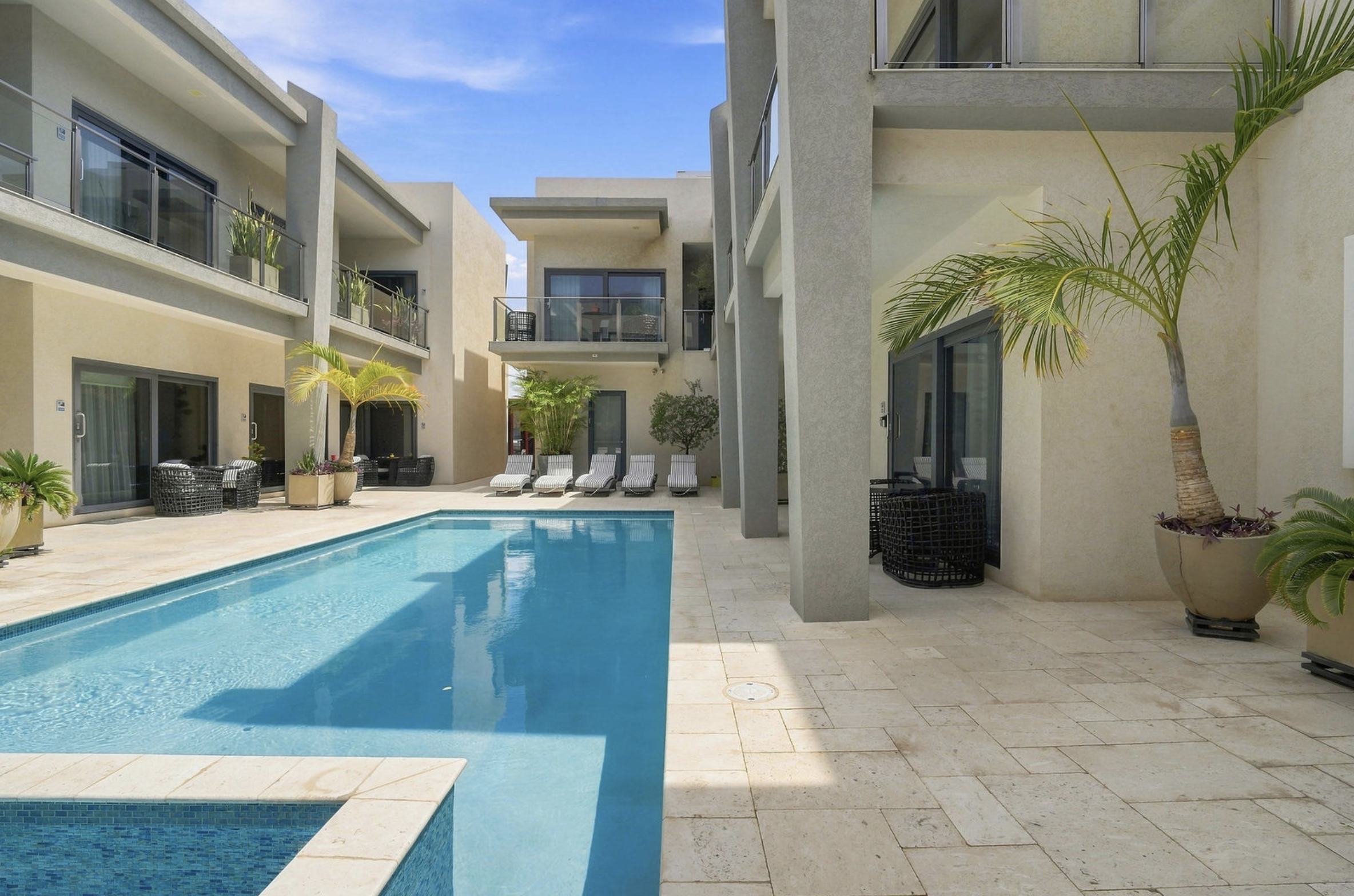 Modern apartment complex with a swimming pool, poolside lounge chairs, and potted palm trees, under a clear blue sky.