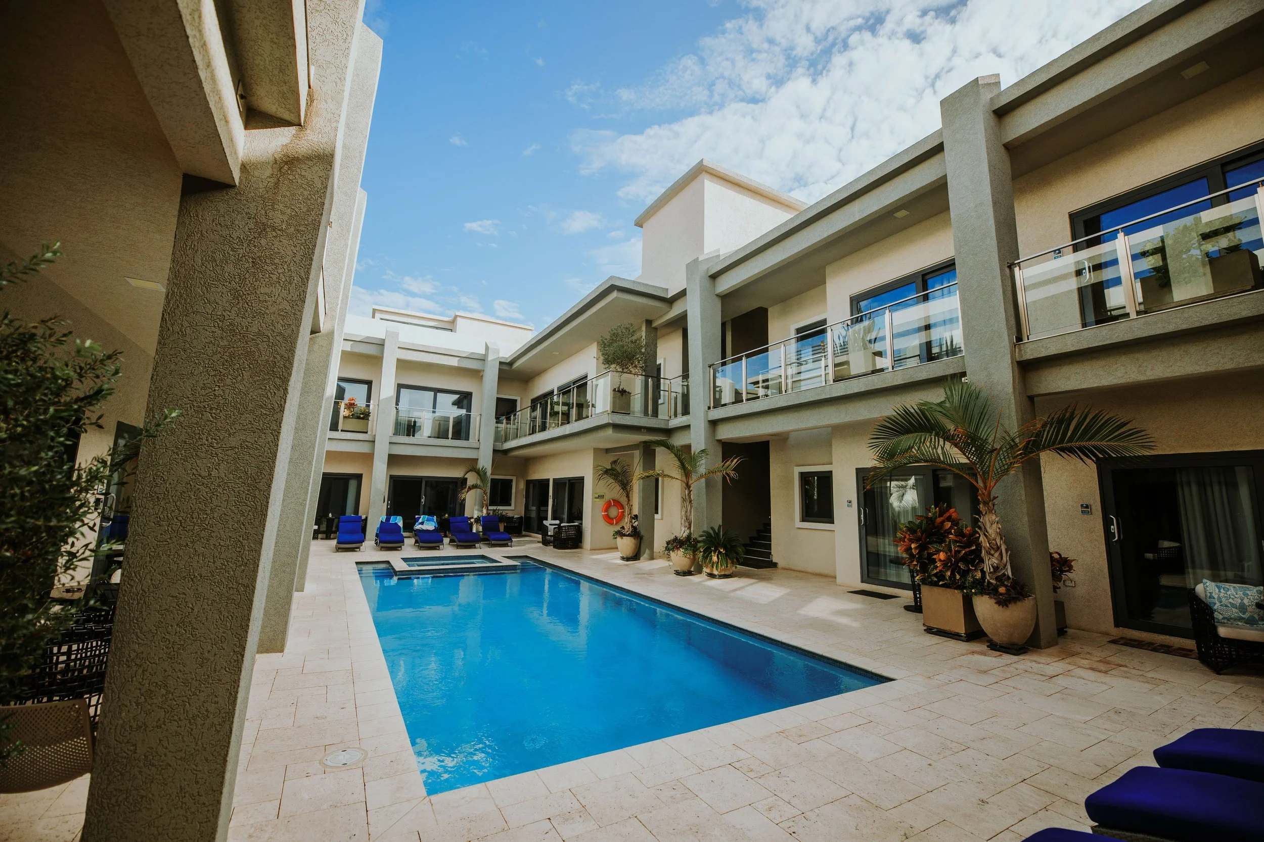 A modern apartment complex with a rectangular swimming pool surrounded by lounge chairs, potted plants, and balconies overlooking the pool area on a clear day with blue skies.