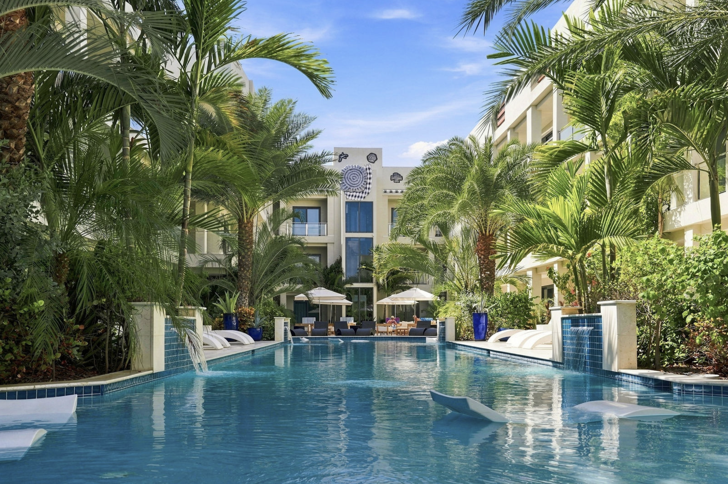 Swimming pool with lounge chairs and umbrellas surrounded by lush palm trees and modern white buildings with balconies under a blue sky with clouds.