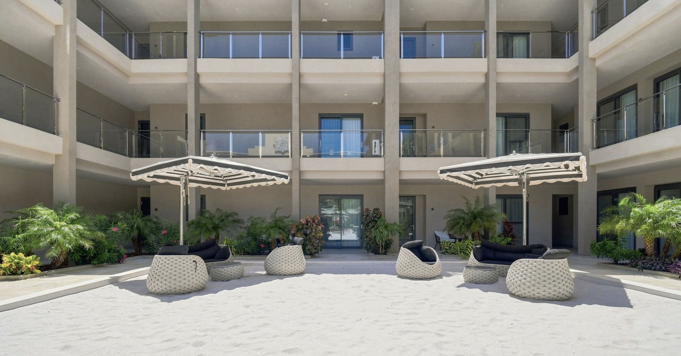 Outdoor courtyard of a modern hotel with lounge chairs, umbrellas, and lush greenery in front of a multi-floor building with balconies.