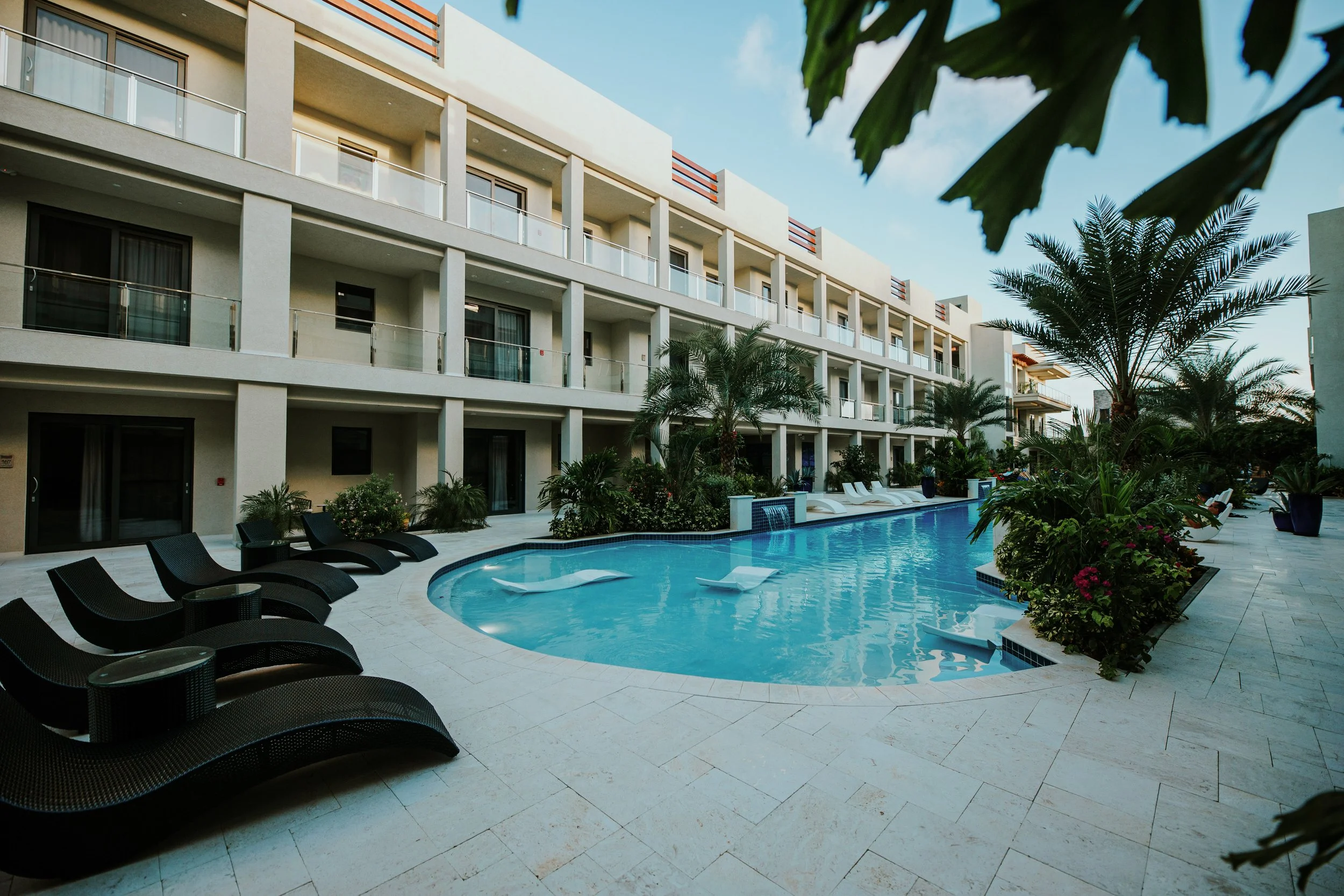 A view of a hotel pool area with lounge chairs, tropical plants, and a multi-story building with balconies in the background.
