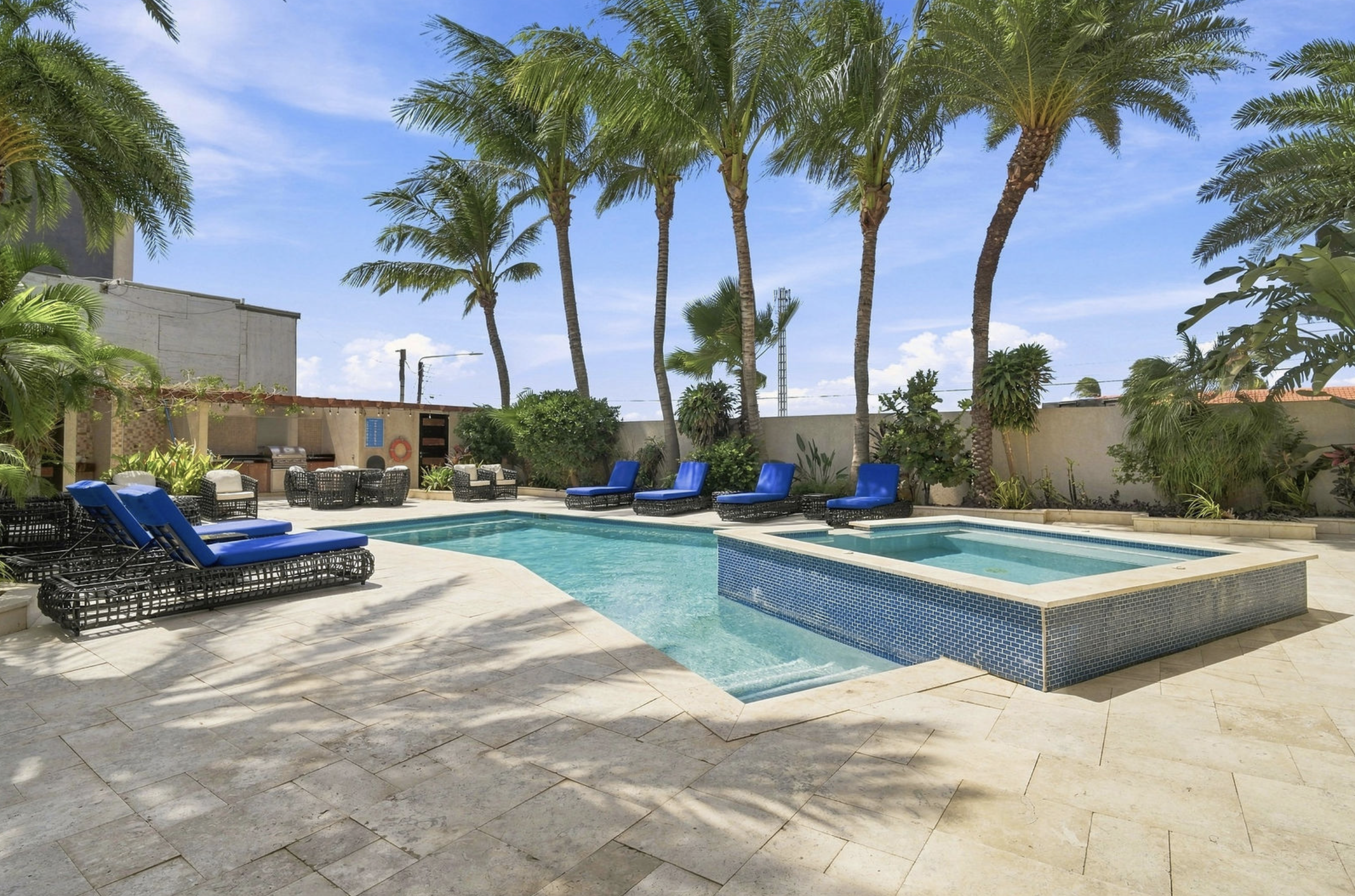 A backyard with a swimming pool surrounded by blue lounge chairs and lush green palm trees under a partly cloudy blue sky.
