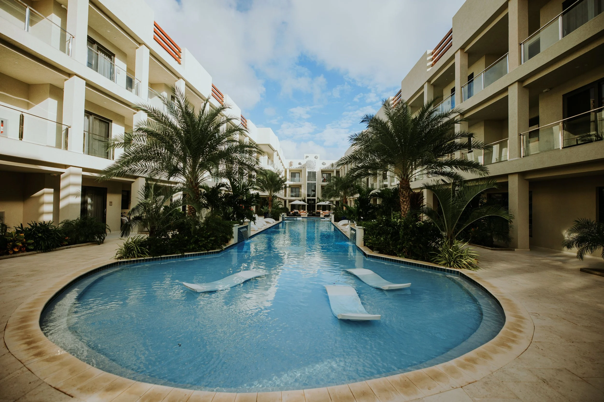 A swimming pool in the courtyard of a hotel with palm trees and lounge chairs, surrounded by modern white buildings under a blue sky with clouds.