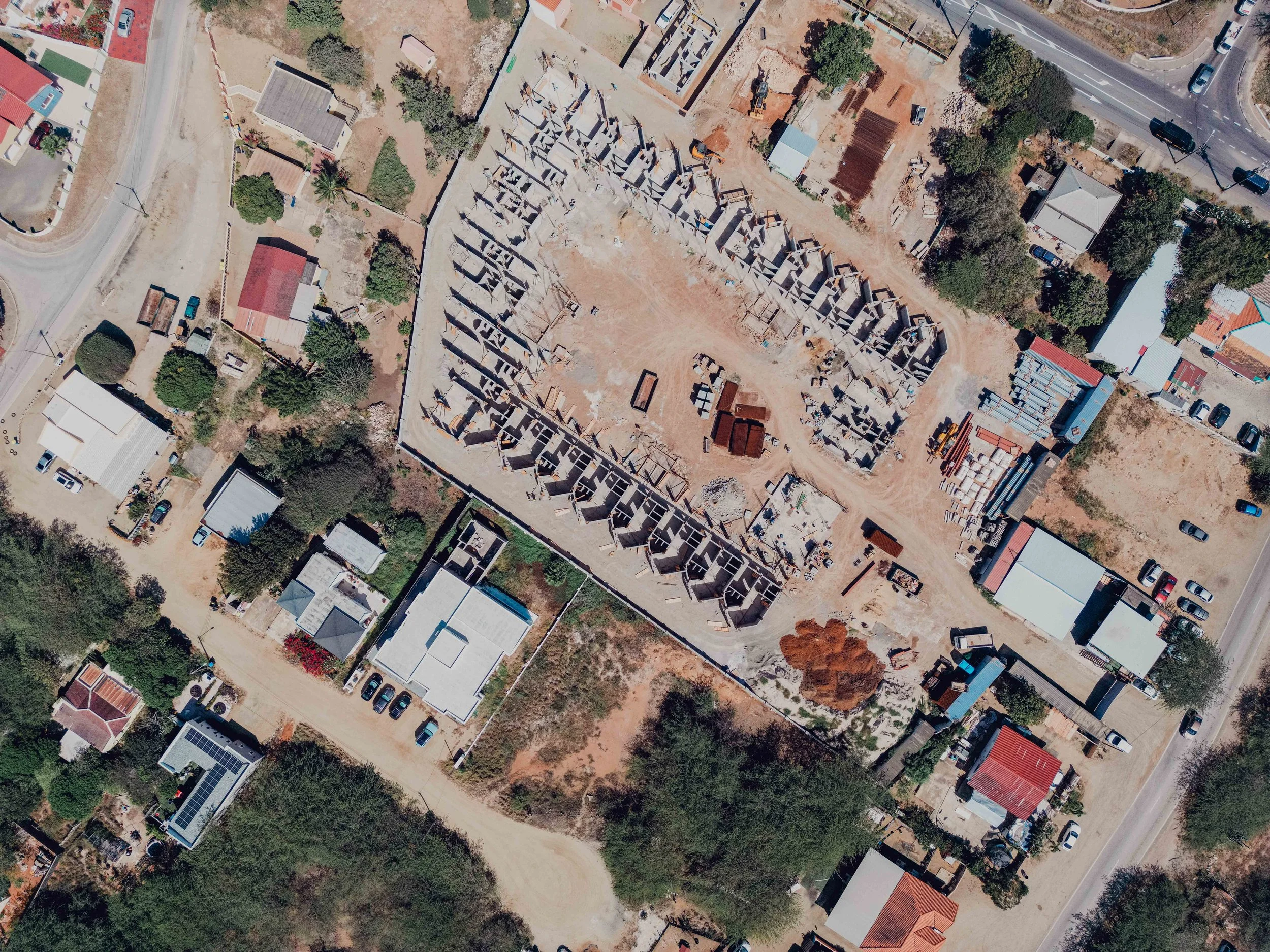 Aerial view of a construction site with multiple incomplete buildings, surrounded by residential houses, roads, and trees.