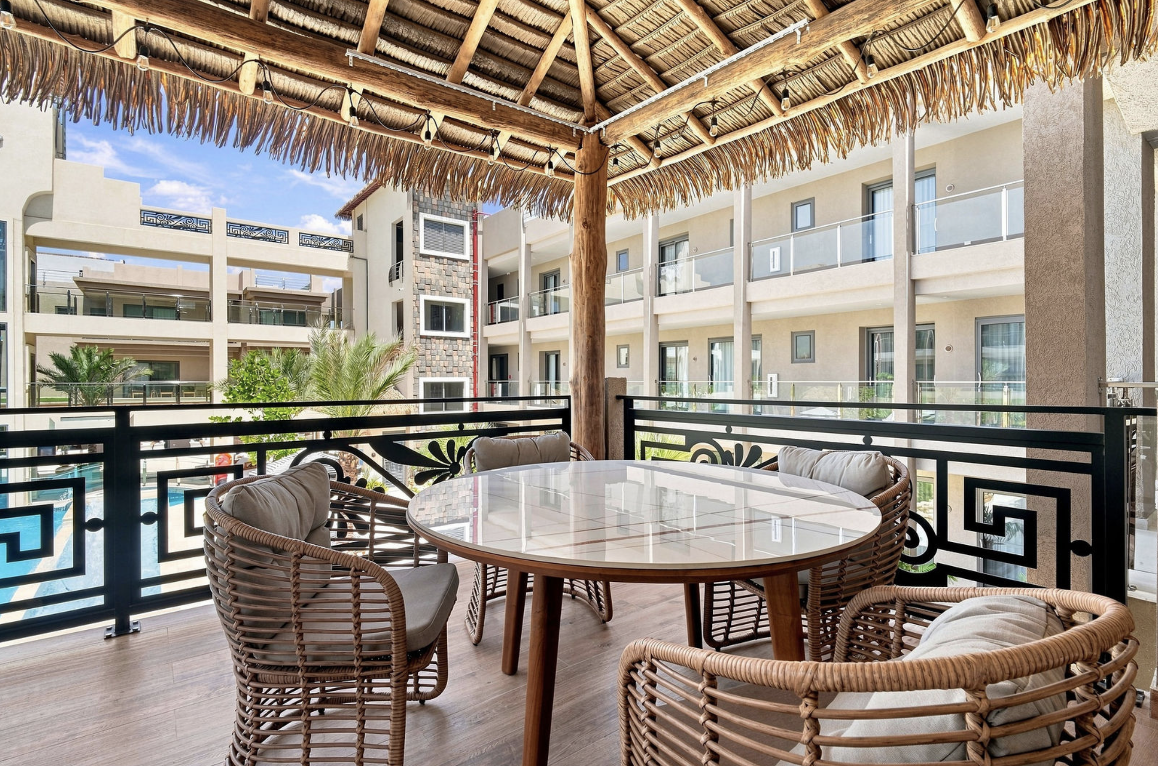 Outdoor patio with round table, wicker chairs with cushions, wooden deck, railing, and a thatched roof. A hotel building's balconies and blue sky are visible in the background.