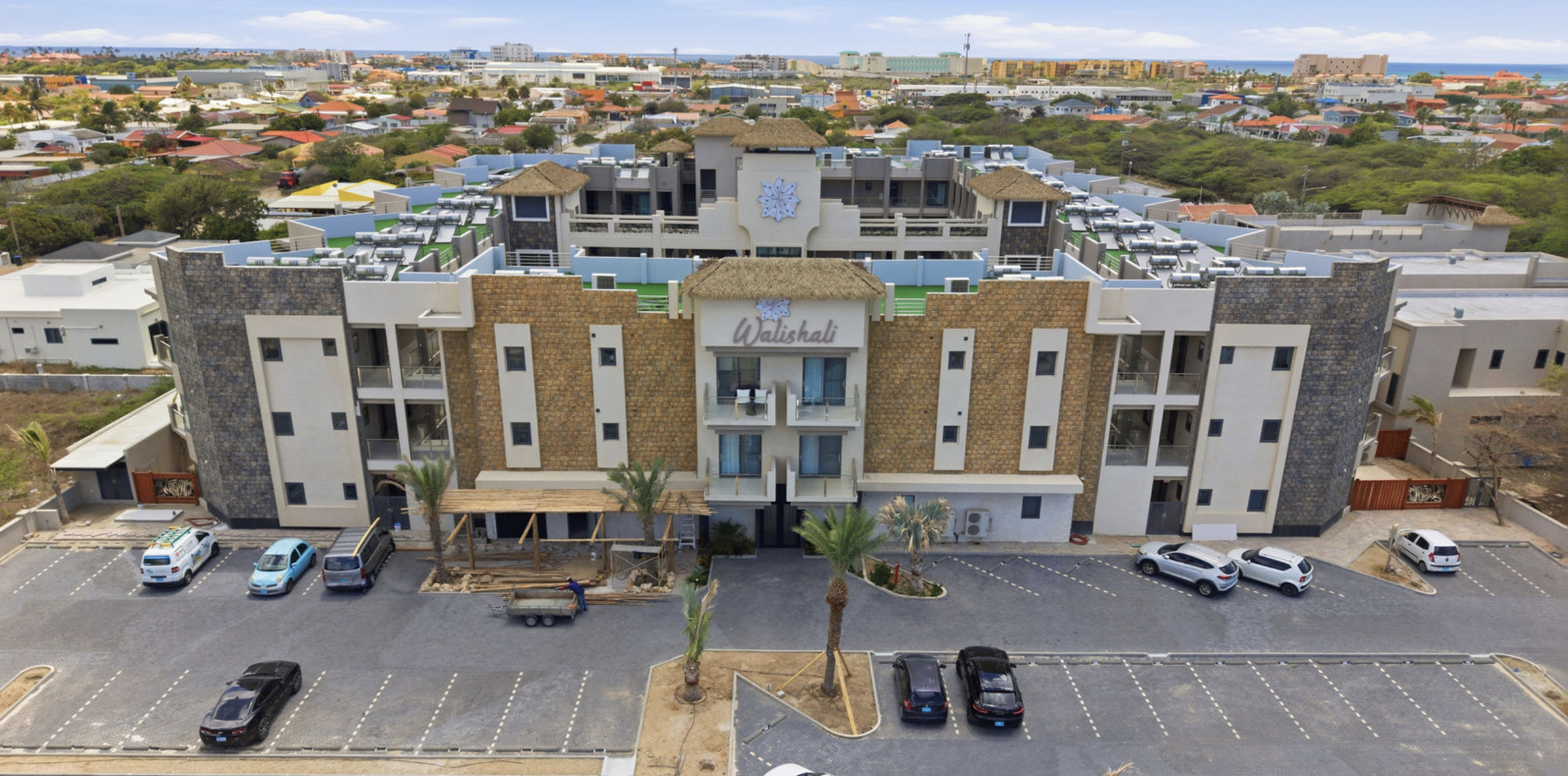 A multi-story hotel building called Walishali with a modern exterior design featuring stone and white wall sections, balconies, and a roof with thatched sections. The hotel has a parking lot in front with several parked cars, and palm trees are plant