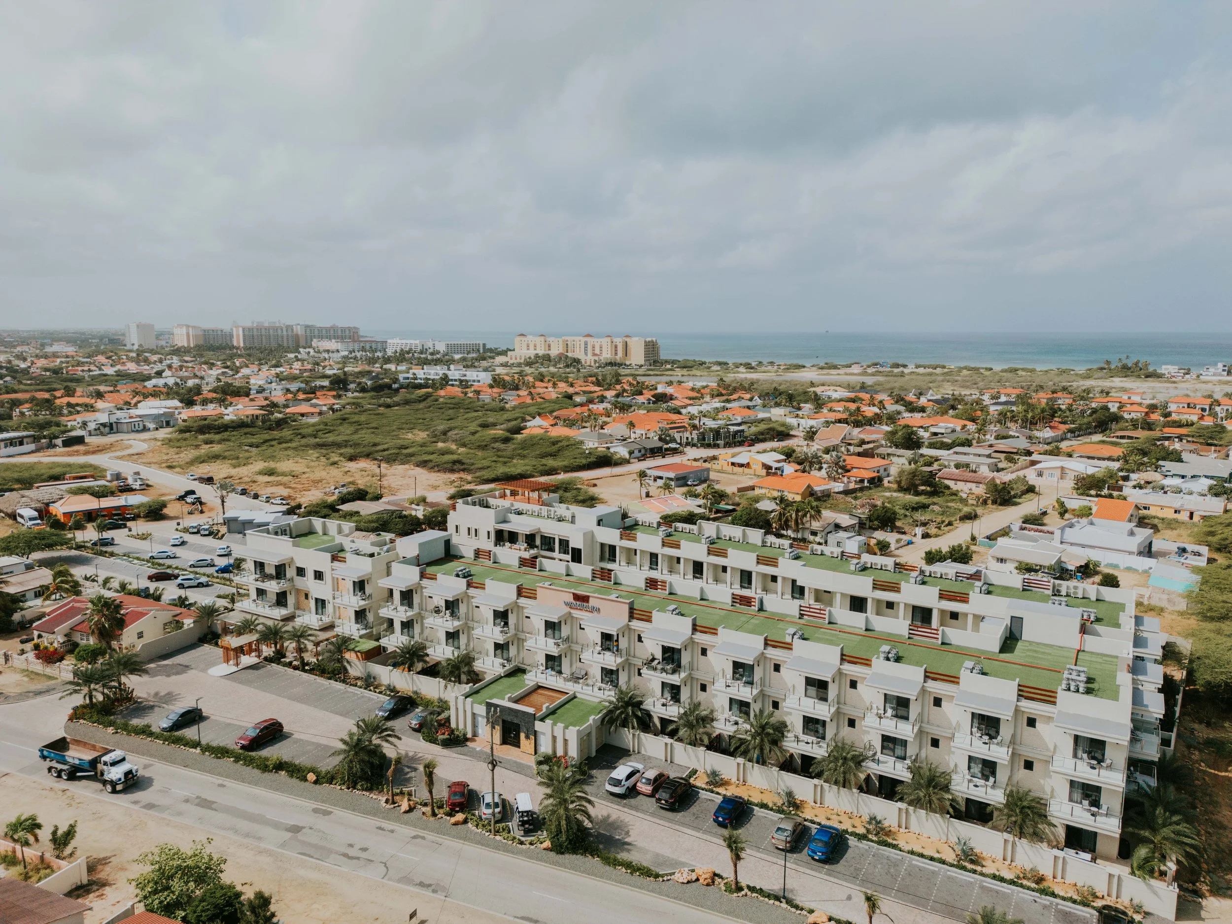 Aerial view of a residential area near a beach with modern white apartment buildings, parked cars, palm trees, and an ocean in the background under a cloudy sky.