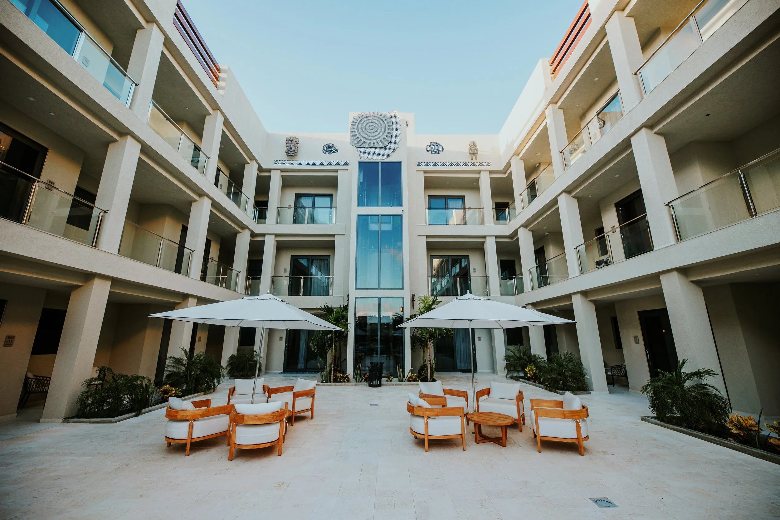 A modern four-story apartment building with balconies and glass railings surrounds a central courtyard with lounge chairs, umbrellas, and tropical plants.