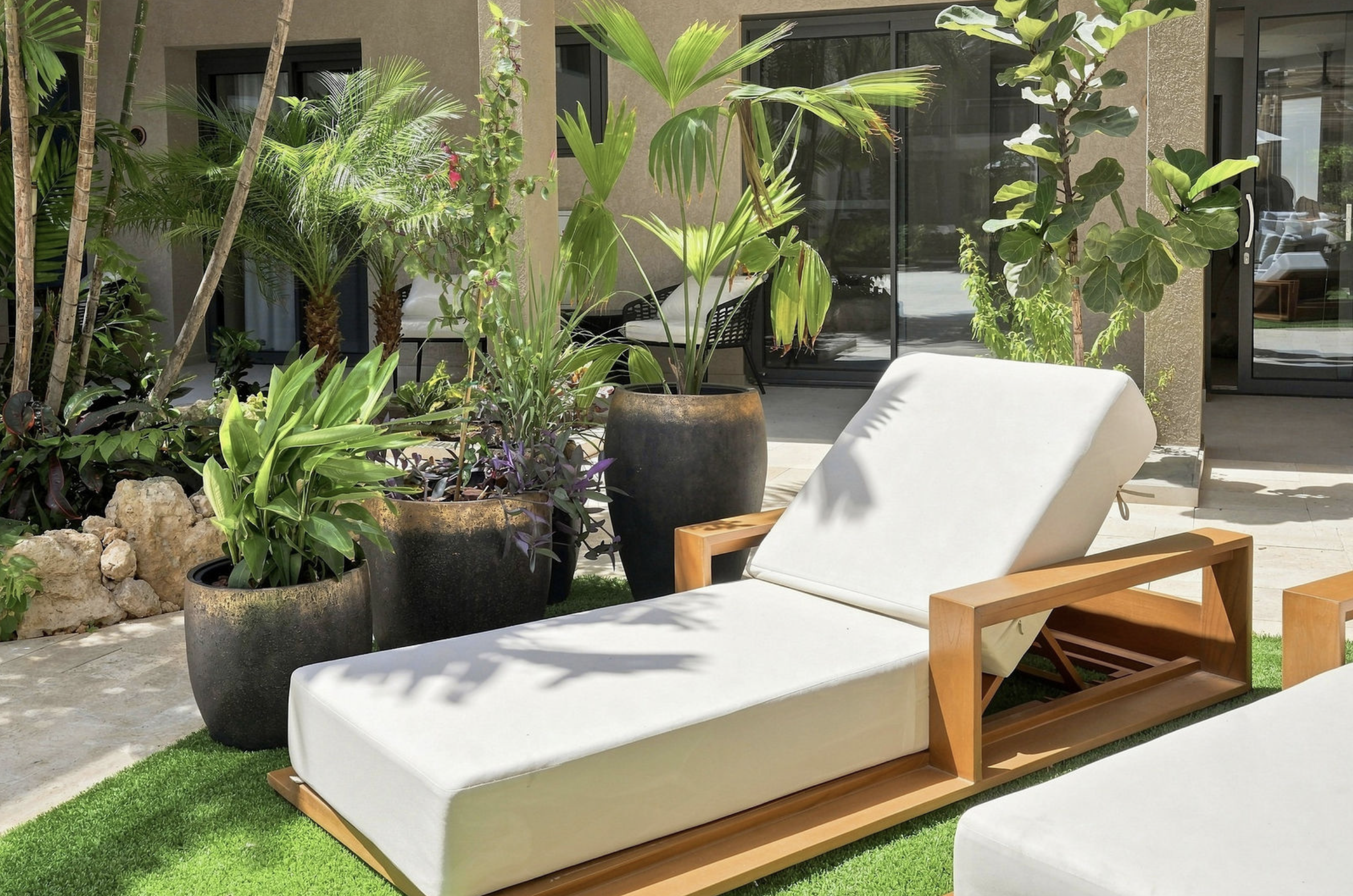 Outdoor patio with white lounge chair with wooden frame, surrounded by potted tropical plants and trees, with glass windows and a beige exterior wall in the background.