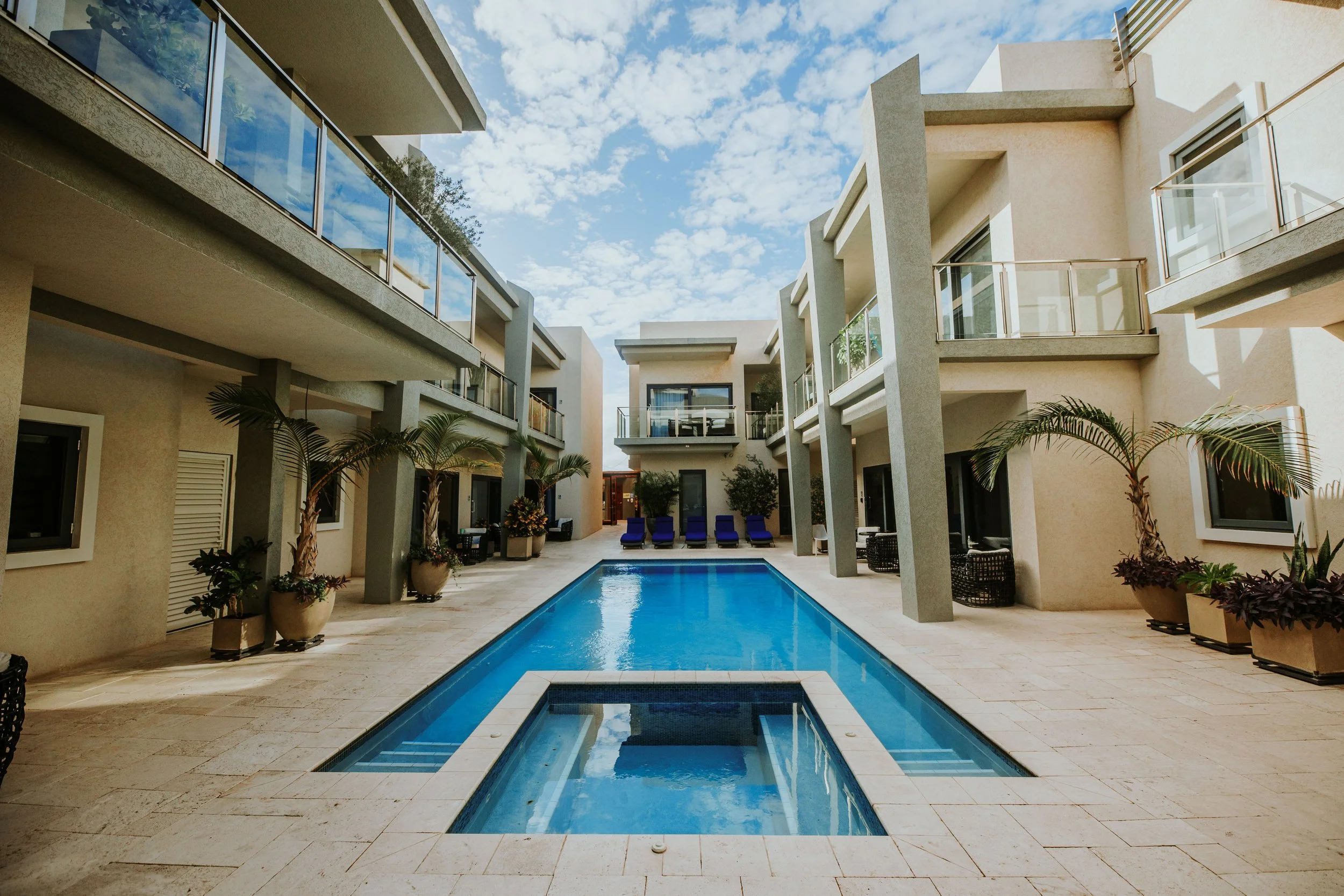 A modern apartment complex featuring a central rectangular swimming pool with lounge chairs at one end, surrounded by beige tiled patio, potted palm plants, and multi-story buildings with balconies and glass railings under a partly cloudy sky.