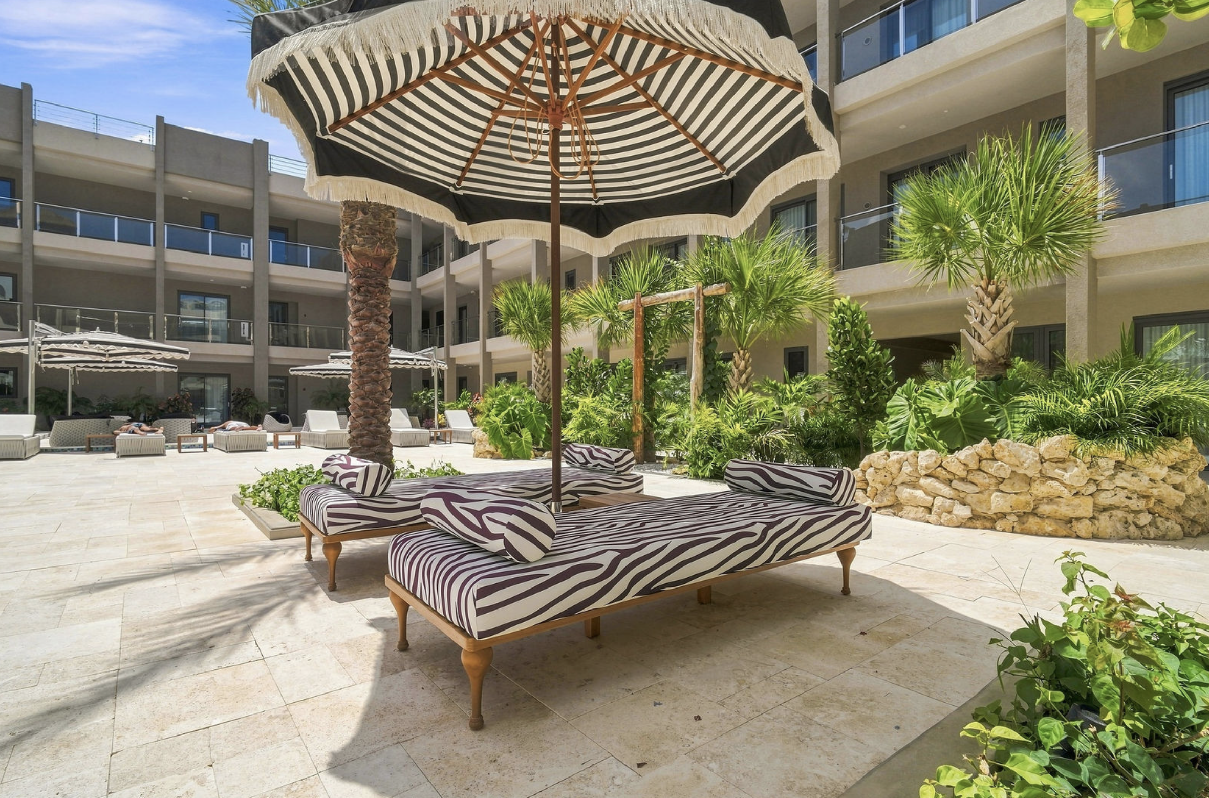 Outdoor poolside area at a hotel or resort with zebra-patterned lounge chairs, a large black and white striped umbrella, green palm trees, and a multi-story building with balconies in the background.