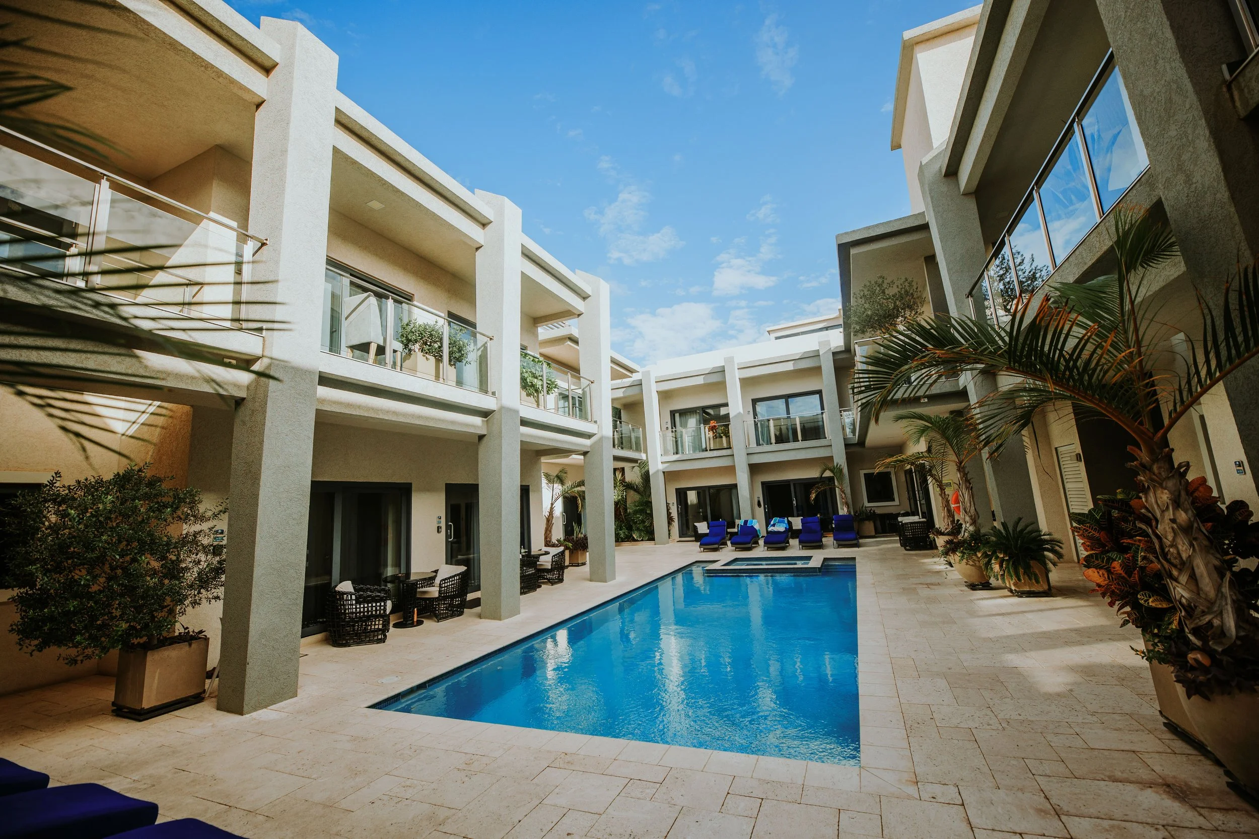 A view of a modern hotel or apartment complex courtyard with a swimming pool, surrounded by lounge chairs, potted plants, and balconies, under a blue sky with some clouds.
