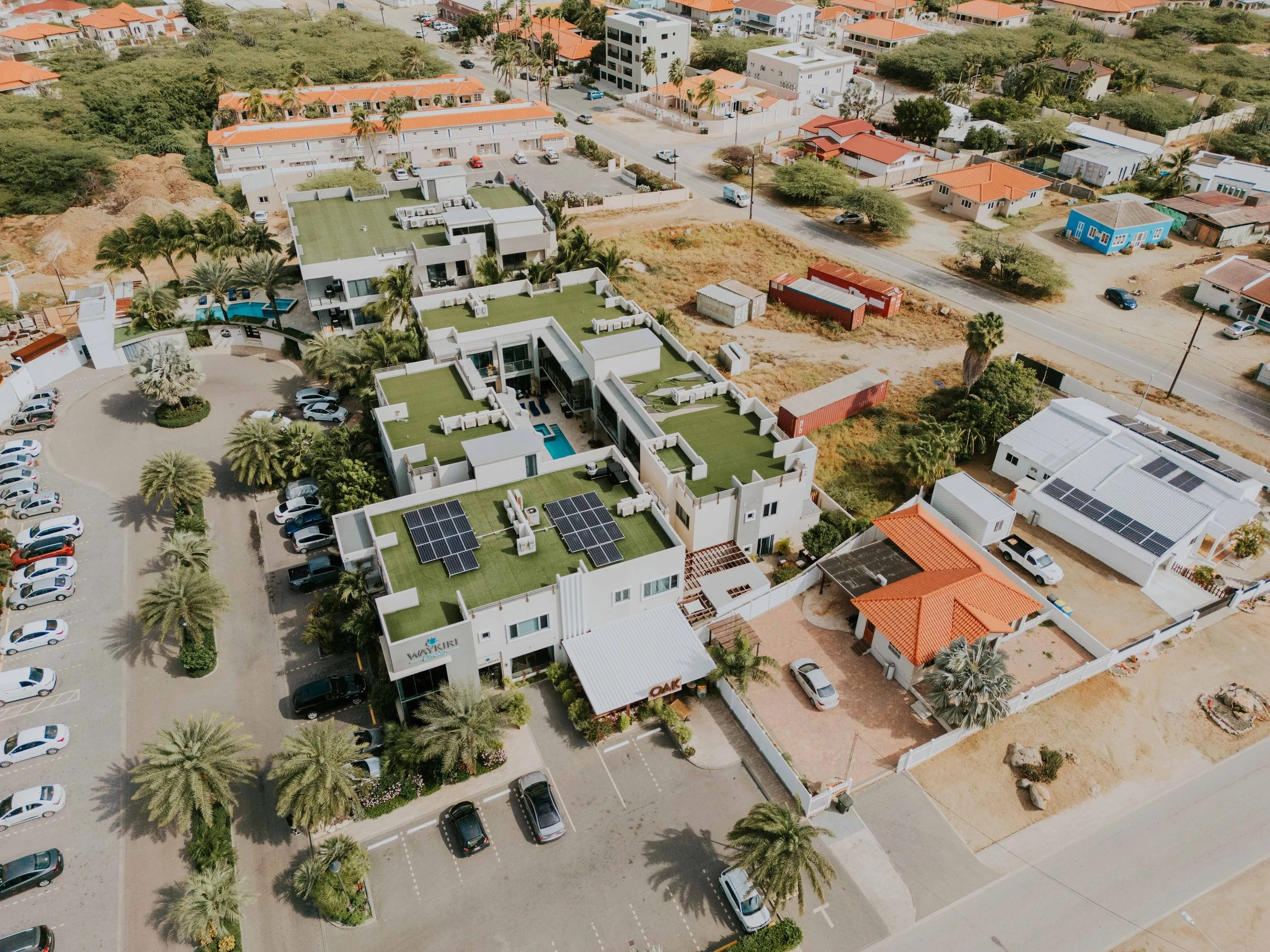 An aerial view of a modern building with solar panels on its green roof, adjacent parking lot, surrounded by palm trees, and neighboring houses with red tile roofs in a suburban area.