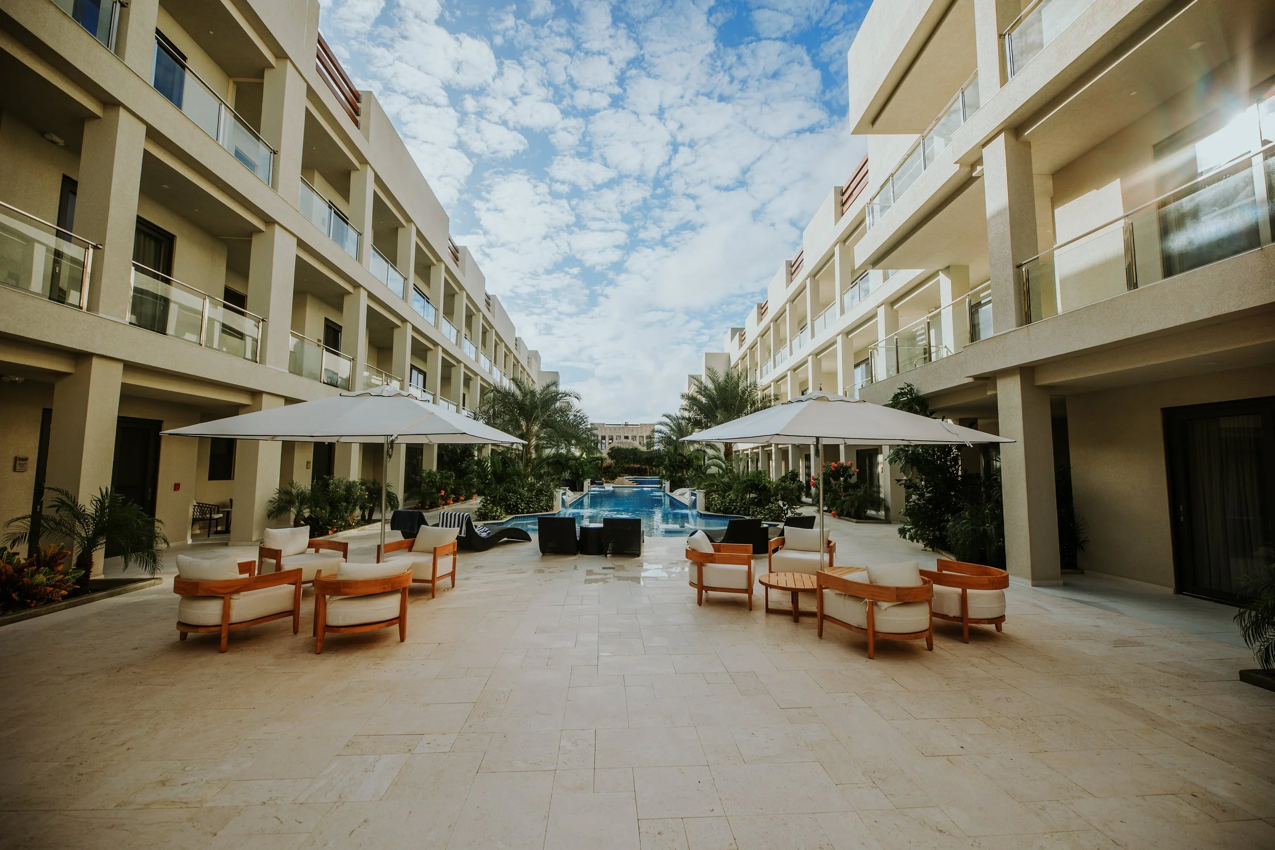 Pool area with seating and umbrellas between modern beige apartment buildings under a partly cloudy sky.