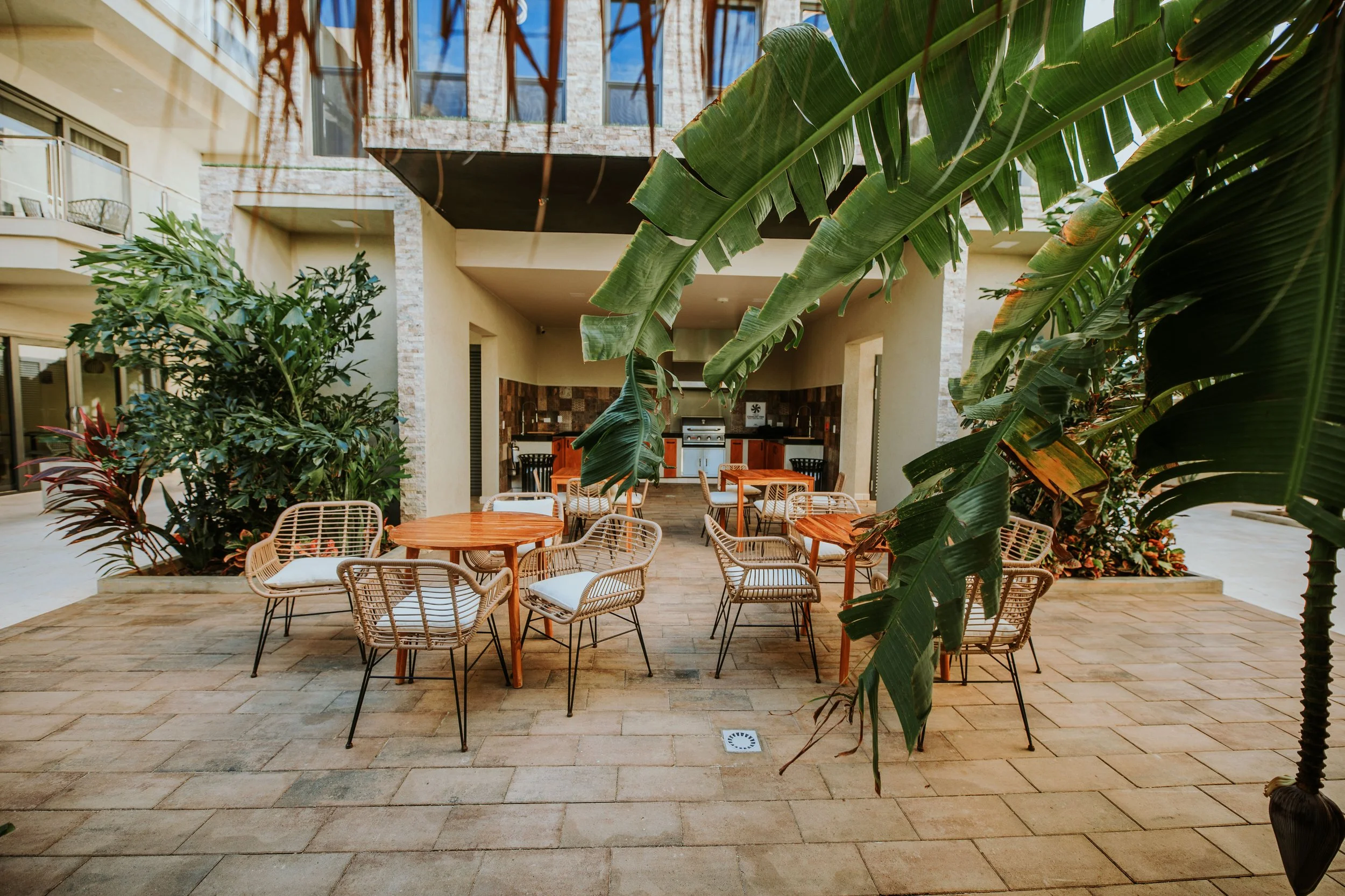 Outdoor patio with wooden tables and wicker chairs, tropical plants, and a built-in outdoor kitchen in the background.