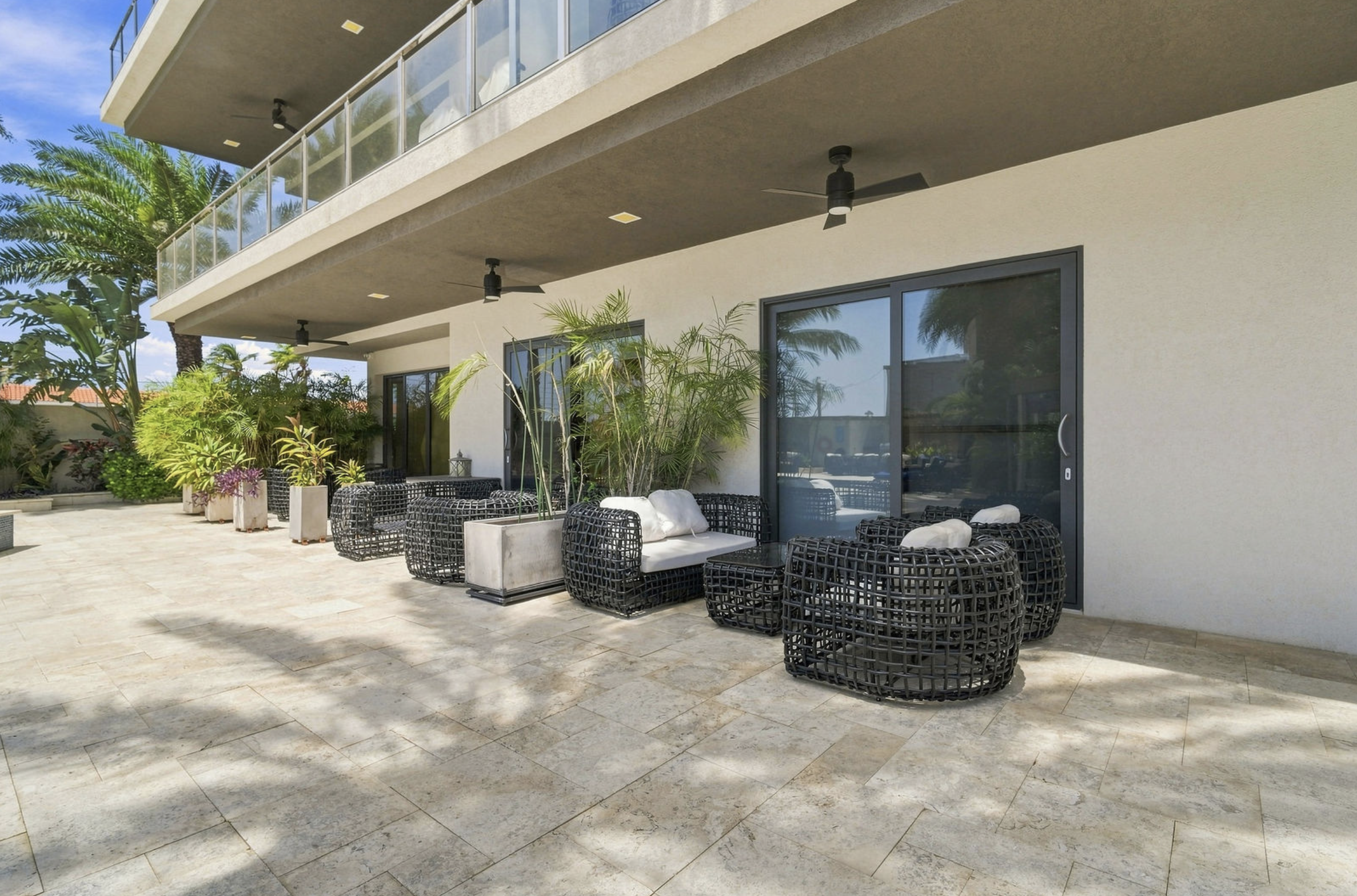 Outdoor patio area with black wicker furniture, potted plants, and glass sliding doors. There are ceiling fans and a balcony above, with tropical trees visible in the background.