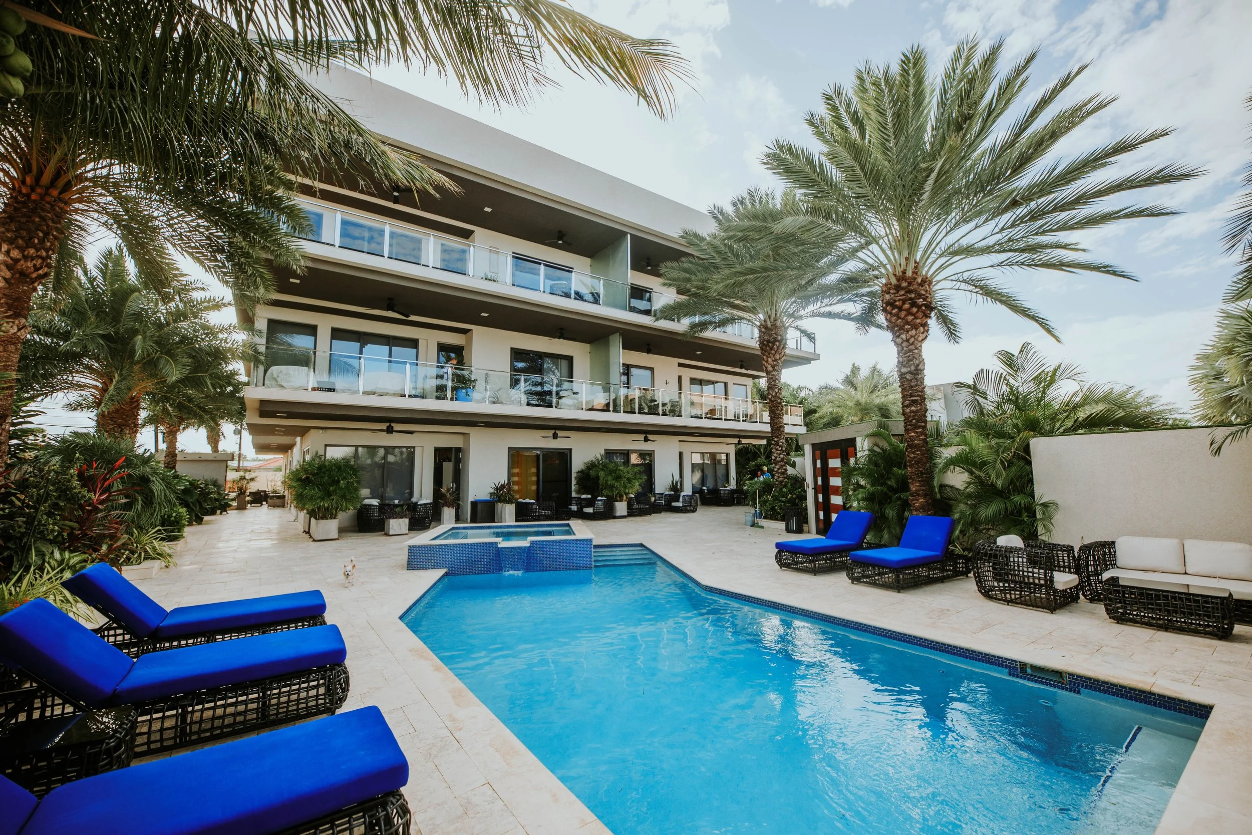 A modern multi-story hotel with balconies, outdoor pool with blue lounge chairs, palm trees, and tropical landscaping in a sunny setting.