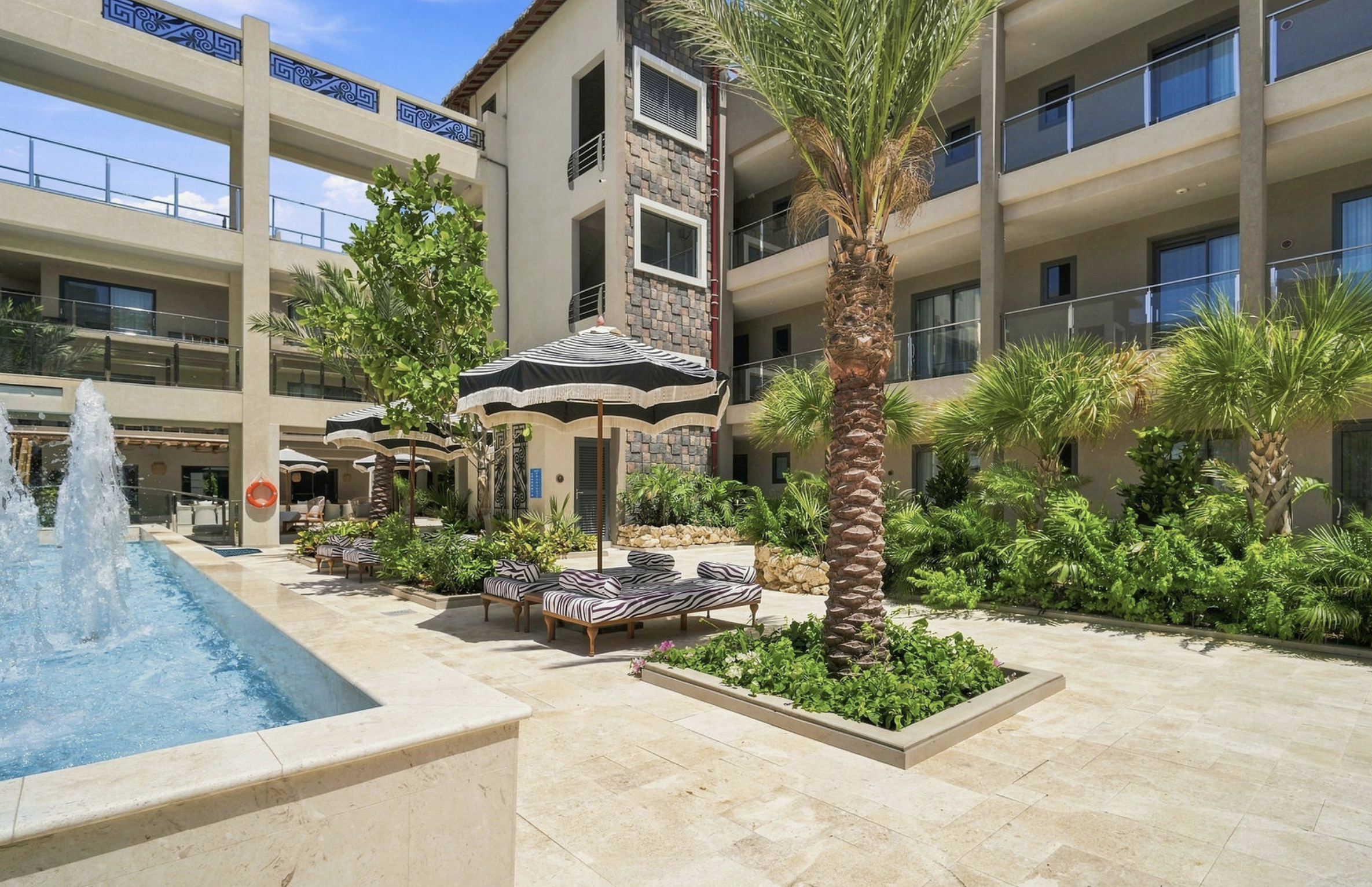 Outdoor pool area of a hotel with lounge chairs with zebra-striped cushions, striped umbrellas, lush greenery, palm trees, a fountain, and a multi-story building with balconies in the background under a blue sky.