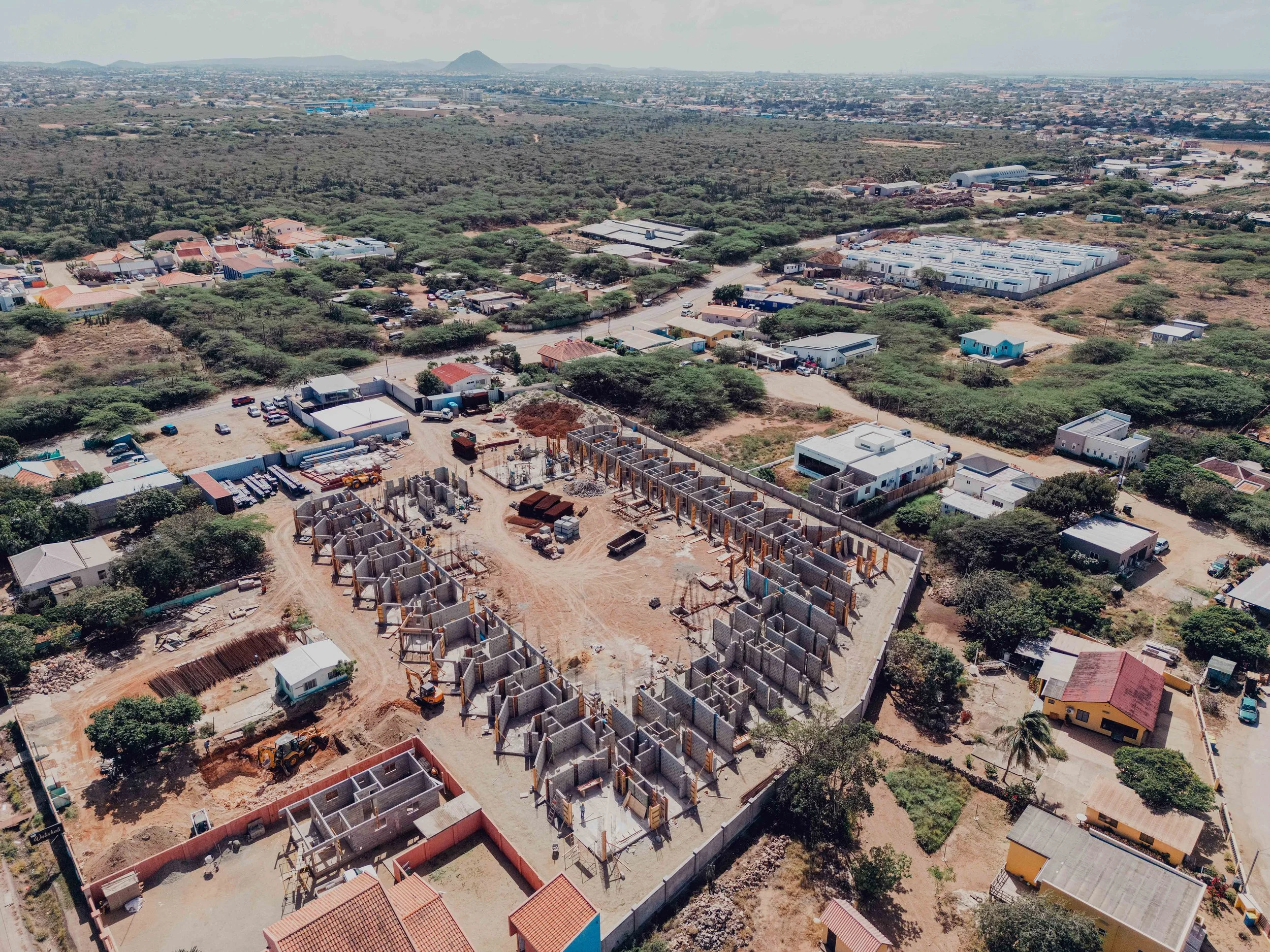 Aerial view of a construction site where a building is being erected, surrounded by houses, small businesses, and greenery in a semi-urban area.