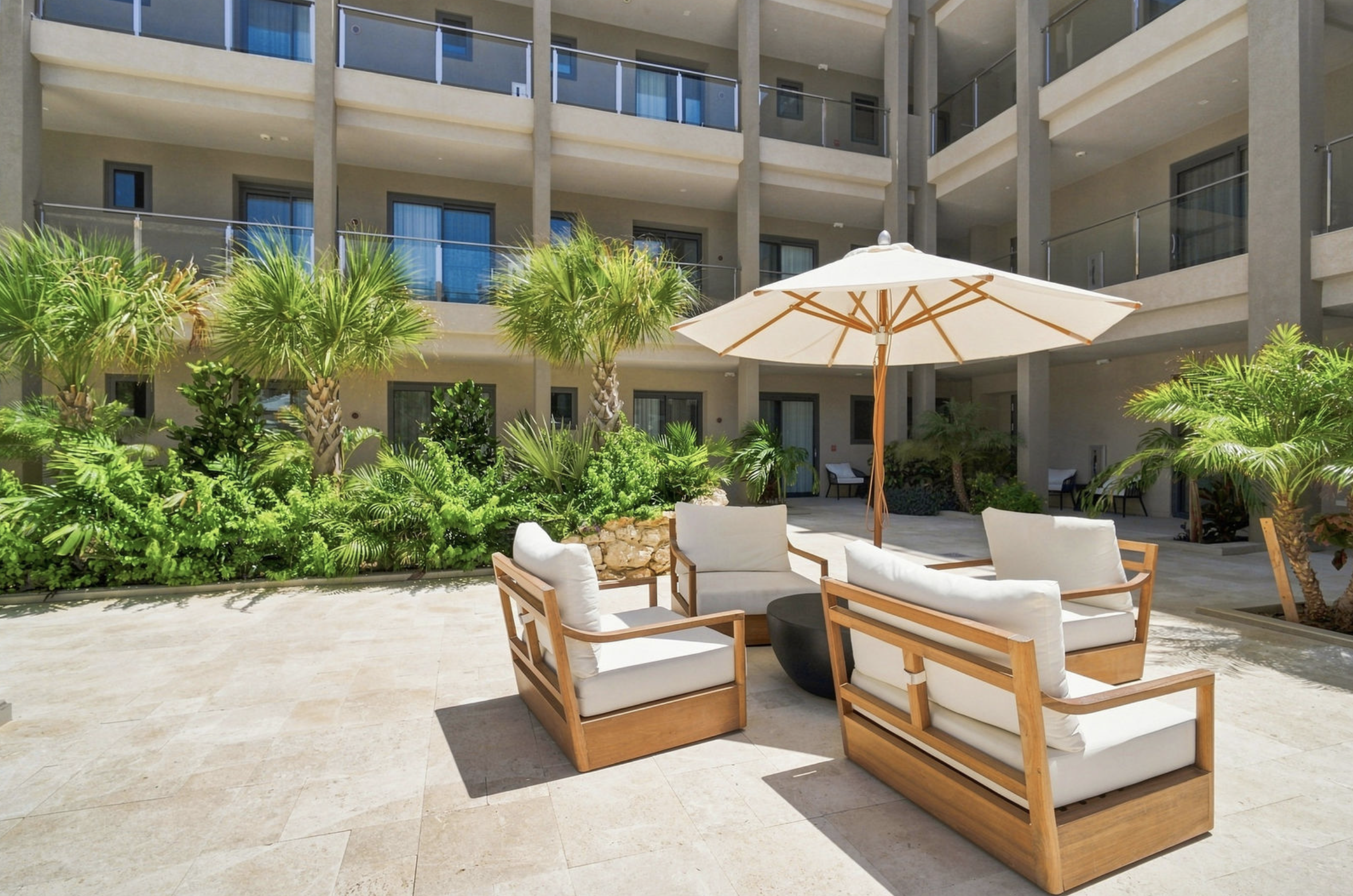 An outdoor poolside lounge area with cushioned wooden furniture, a large umbrella, and lush green plants in front of a modern apartment building with balconies.