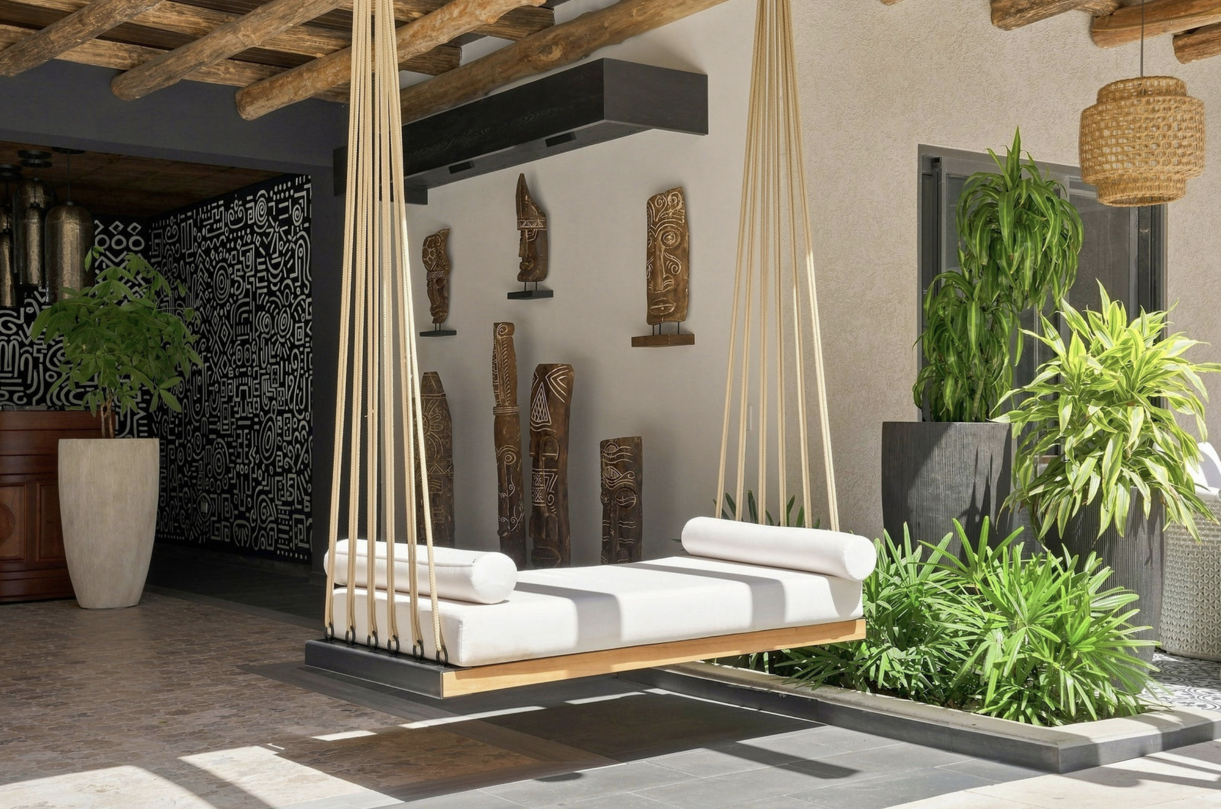 A white cushioned swing with ropes hanging from a wooden ceiling, surrounded by large green potted plants, with tribal art and a black patterned wall in the background.