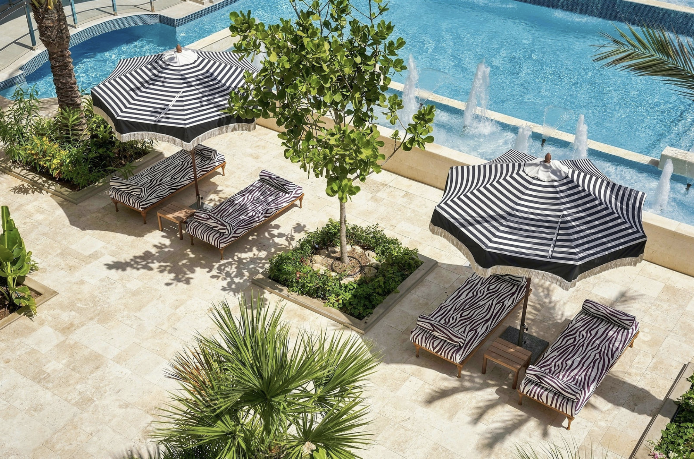 Aerial view of a swimming pool area with striped umbrellas, lounge chairs, potted plants, and a small tree on a tiled surface.