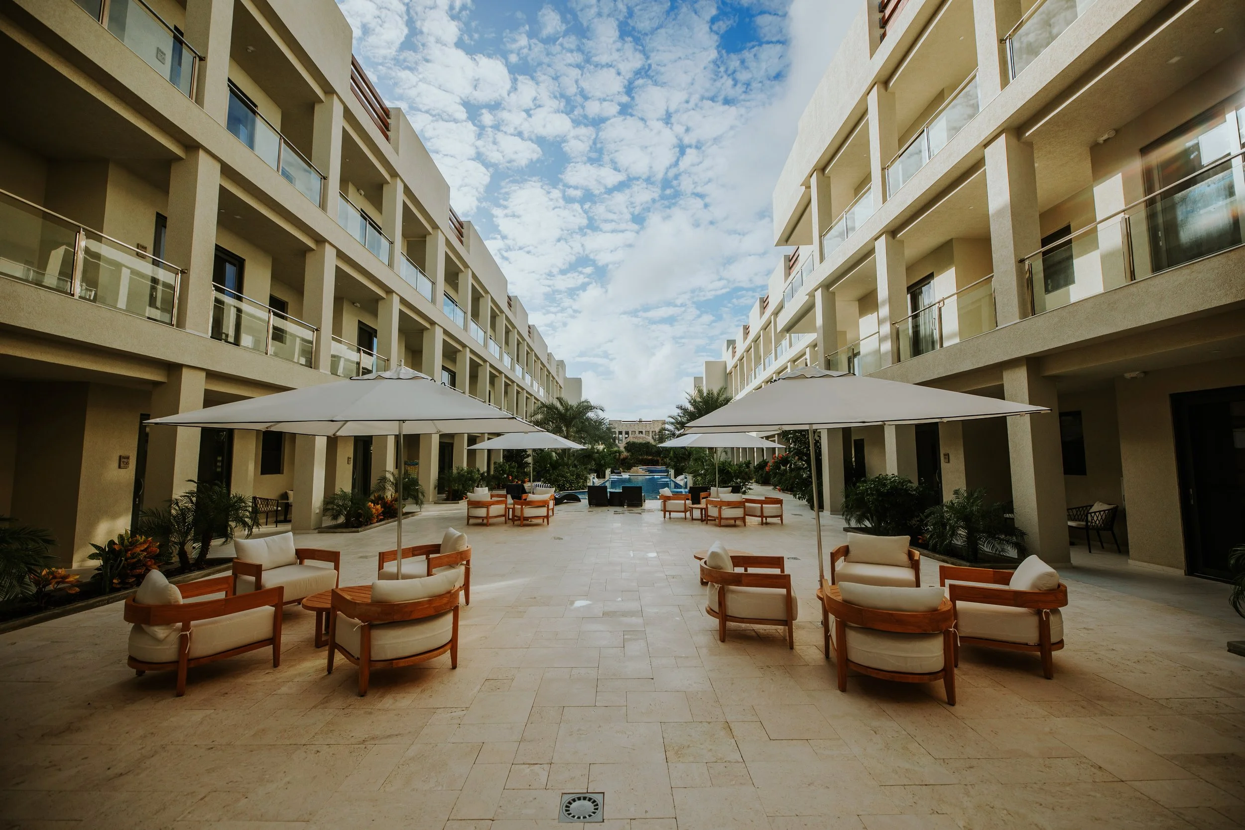 Outdoor patio area with white chairs, tables, umbrellas, and plants, located between beige buildings with balconies, under a partly cloudy sky.