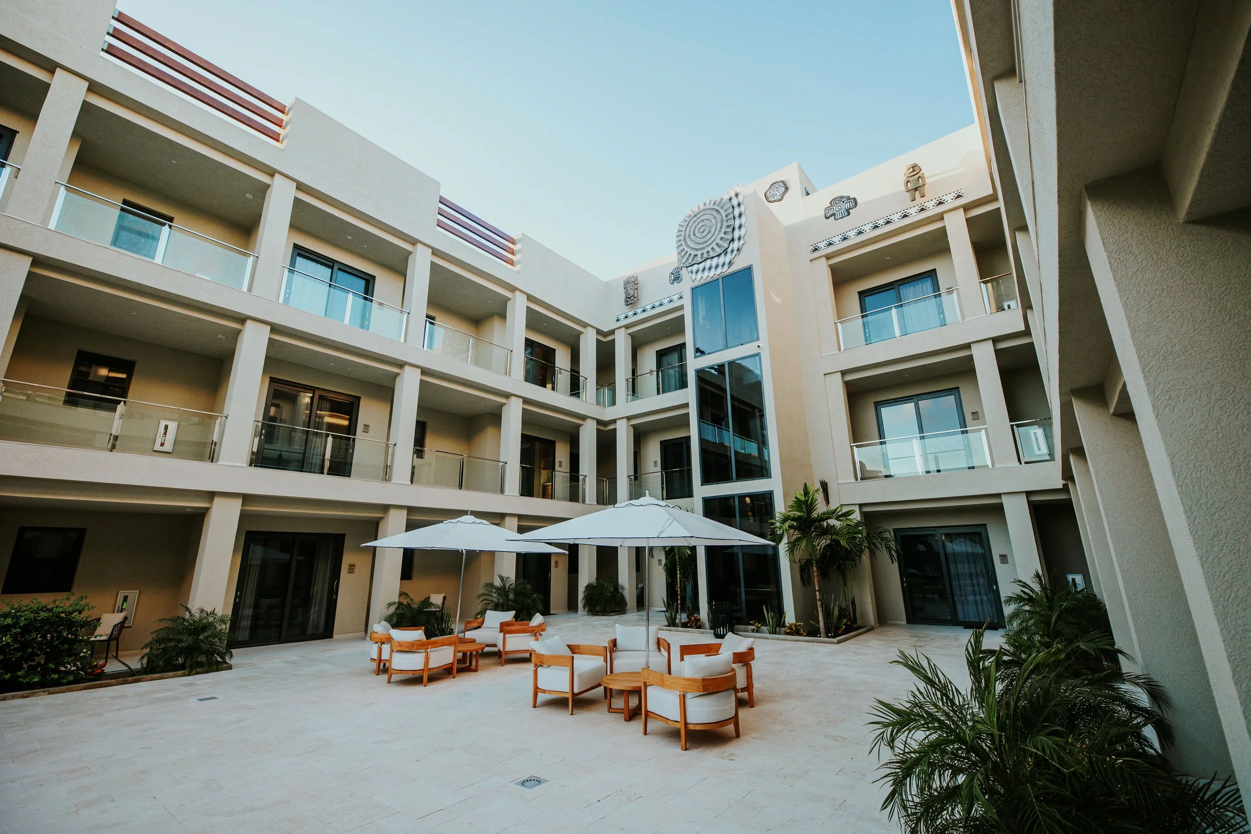 A modern hotel courtyard with outdoor seating, umbrellas, and potted plants, surrounded by a four-story building with balconies.