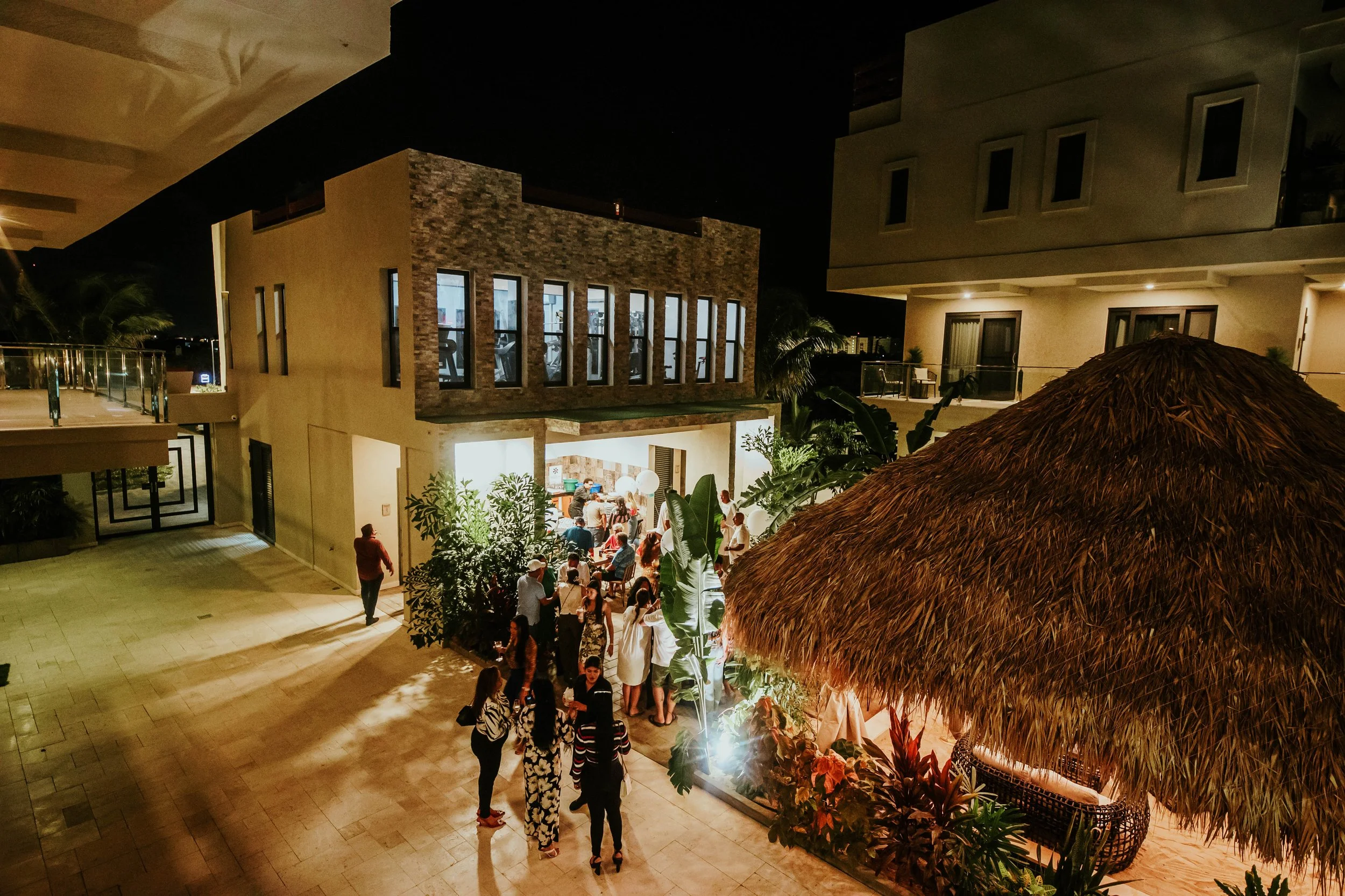 Nighttime scene of a modern hotel or resort with people gathering outside a lit-up patio area, featuring a thatched roof gazebo, lush plants, and multi-story buildings with balconies.