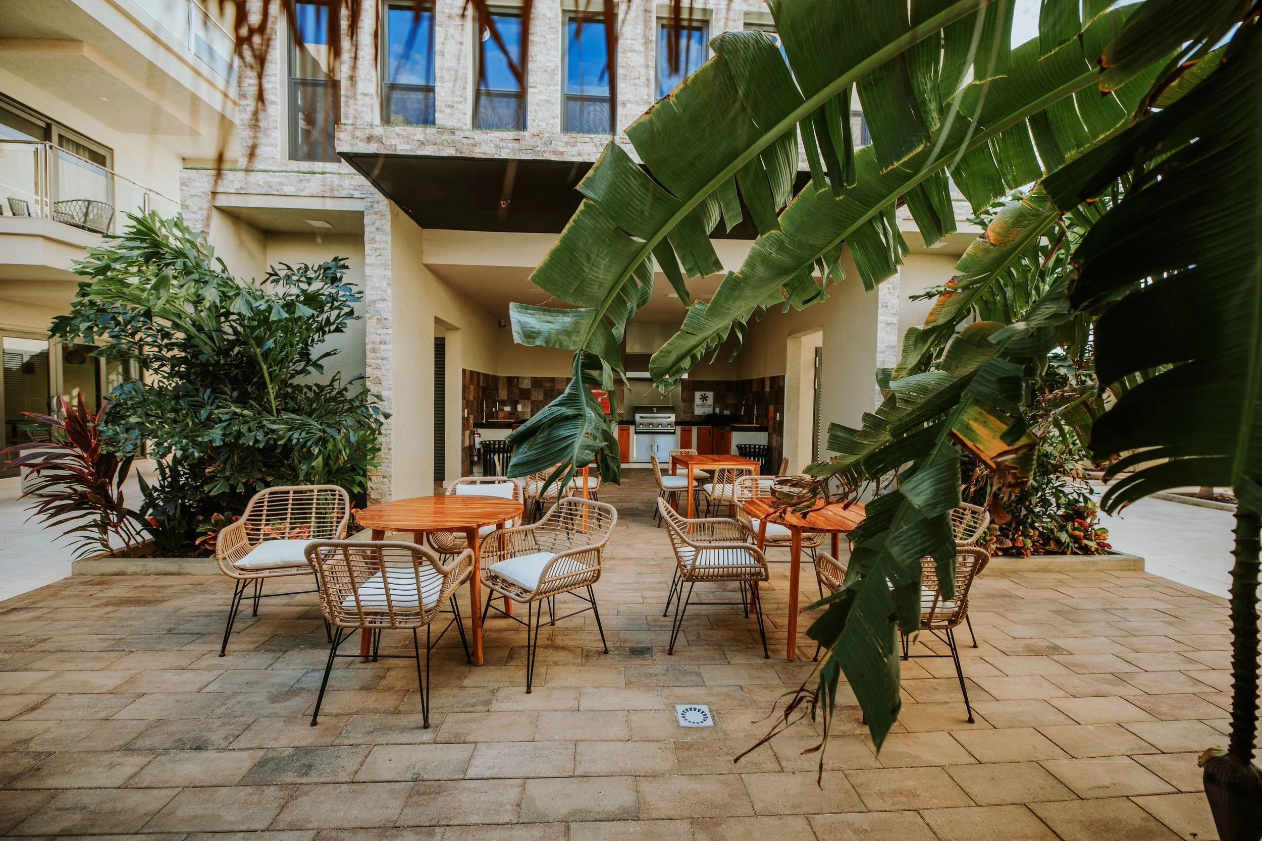 Outdoor dining area with round wooden tables and wicker chairs with cushions, surrounded by tropical plants, in front of a modern building with balconies, and a small outdoor kitchen visible at the back.
