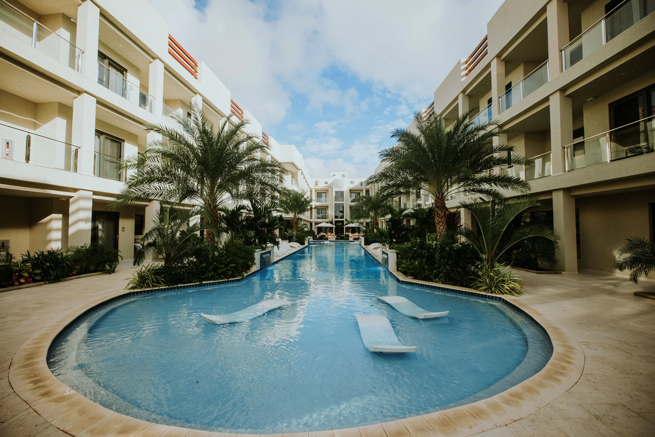 View of a luxurious swimming pool at a modern hotel, surrounded by palm trees and multi-story white buildings with balconies, under a partly cloudy sky.