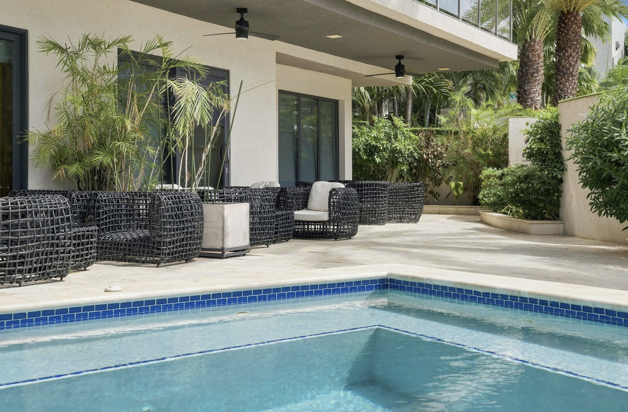 Outdoor pool with a light-colored tiled deck, black wicker lounge chairs with white cushions, a white planter, green tropical plants, a balcony with glass railing, and two black ceiling fans.