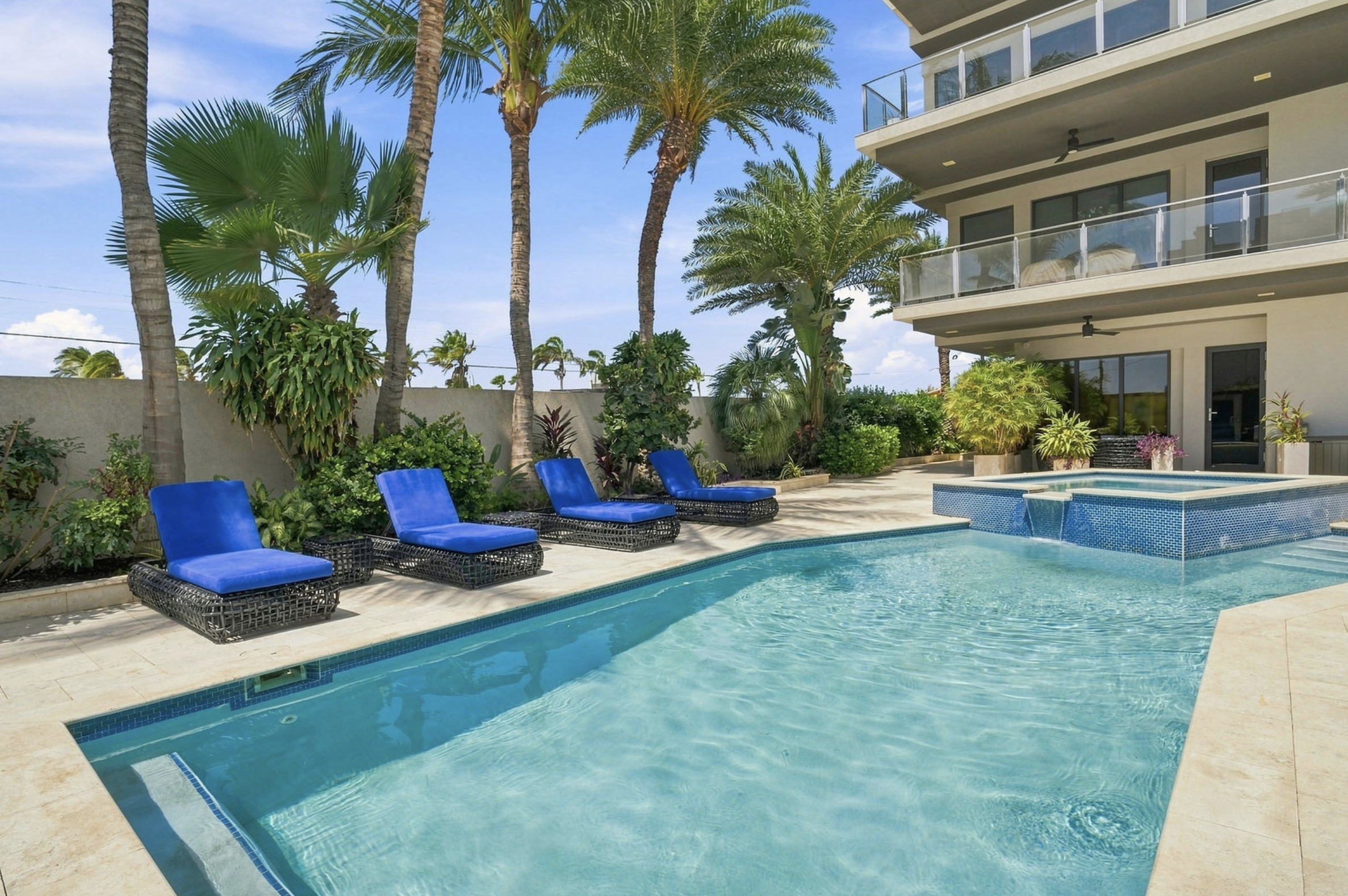 A backyard pool area with blue lounge chairs, lush tropical plants, tall palm trees, and an apartment building with balconies in the background, under a blue sky.