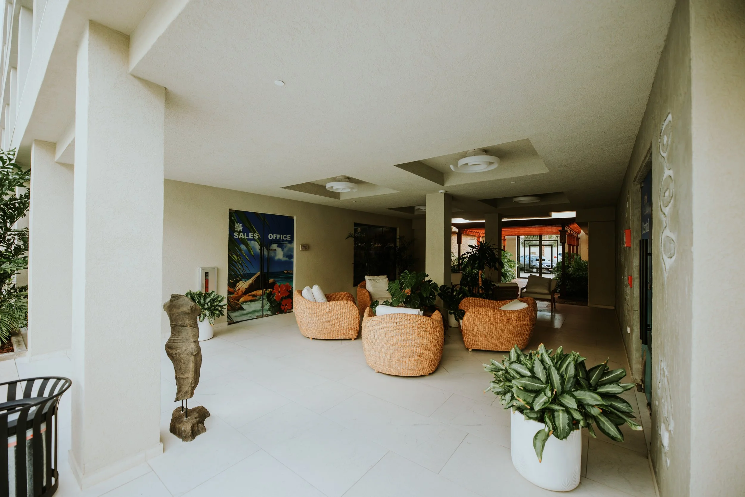Indoor lounge area with wicker chairs, potted plants, and decorative sculpture, near an office and sales area.