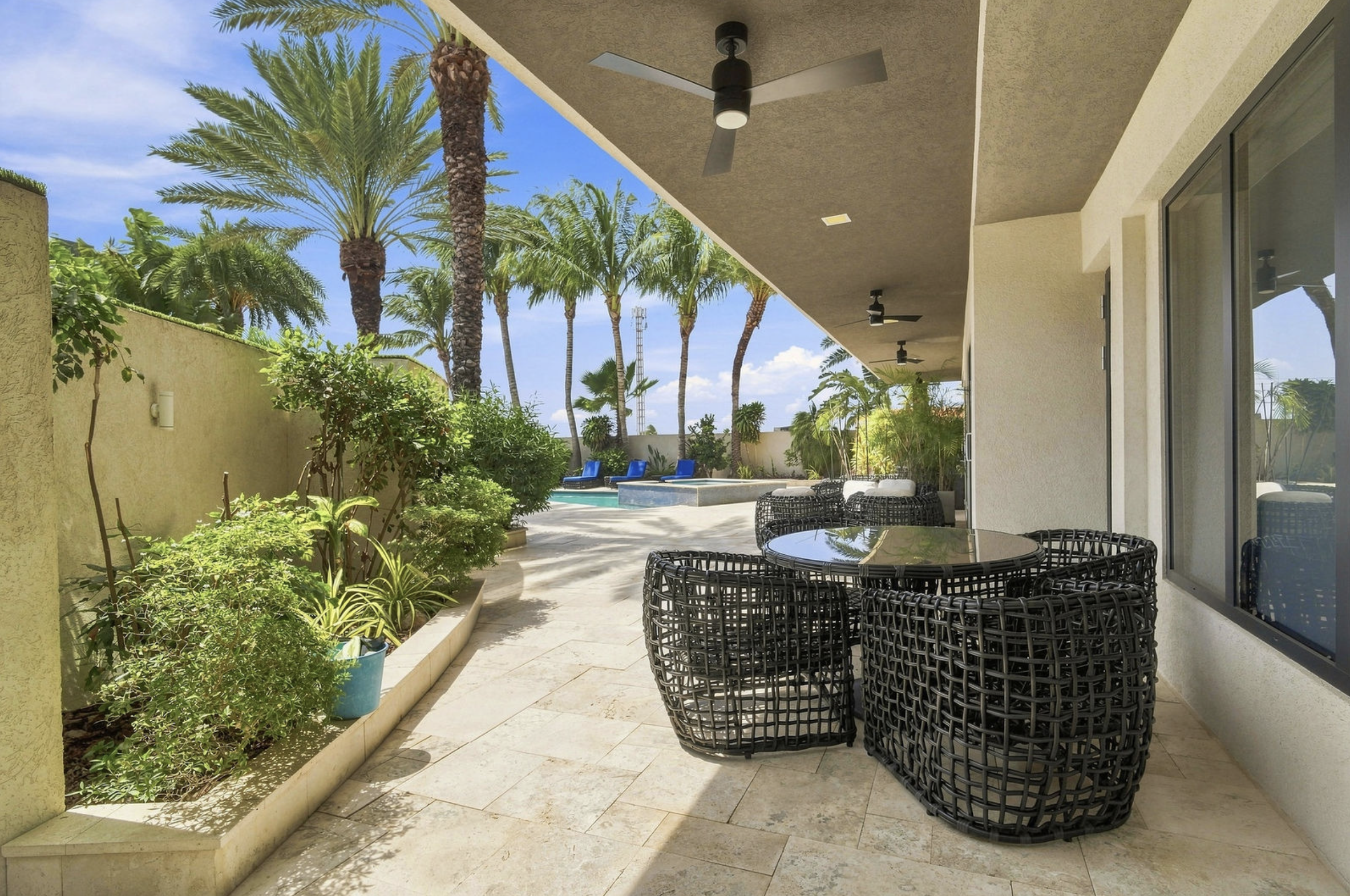 Patio area with black wicker chairs and a glass table, surrounded by lush green plants, with a view of a swimming pool and tall palm trees under a bright blue sky.