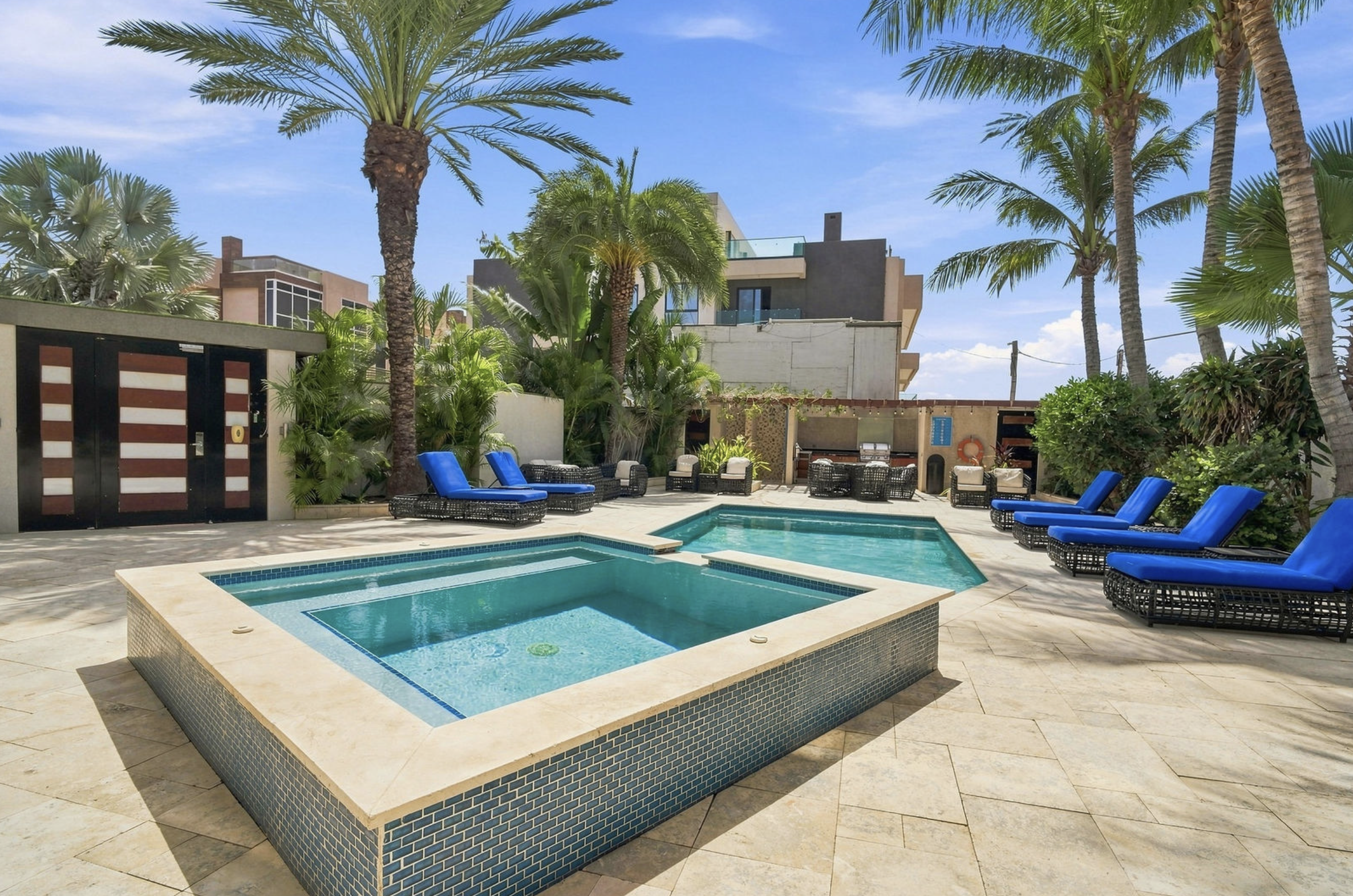 A backyard with a swimming pool, surrounded by palm trees and outdoor seating with blue cushions, overlooking modern house buildings under a blue sky.
