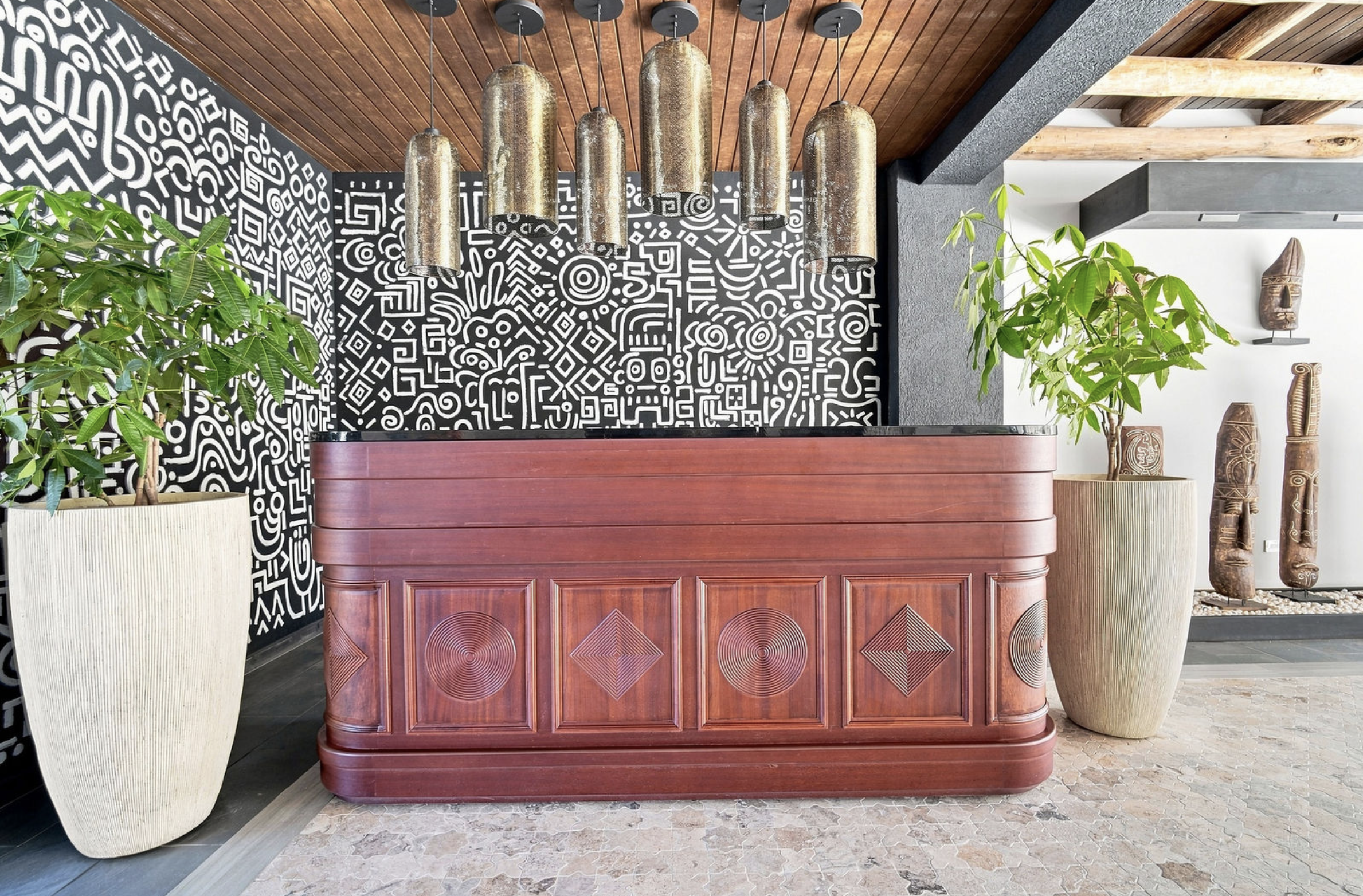 Interior with a wooden reception desk, large black and white geometric patterned wall, and decorative African tribal masks on the wall, with plants in large pots.