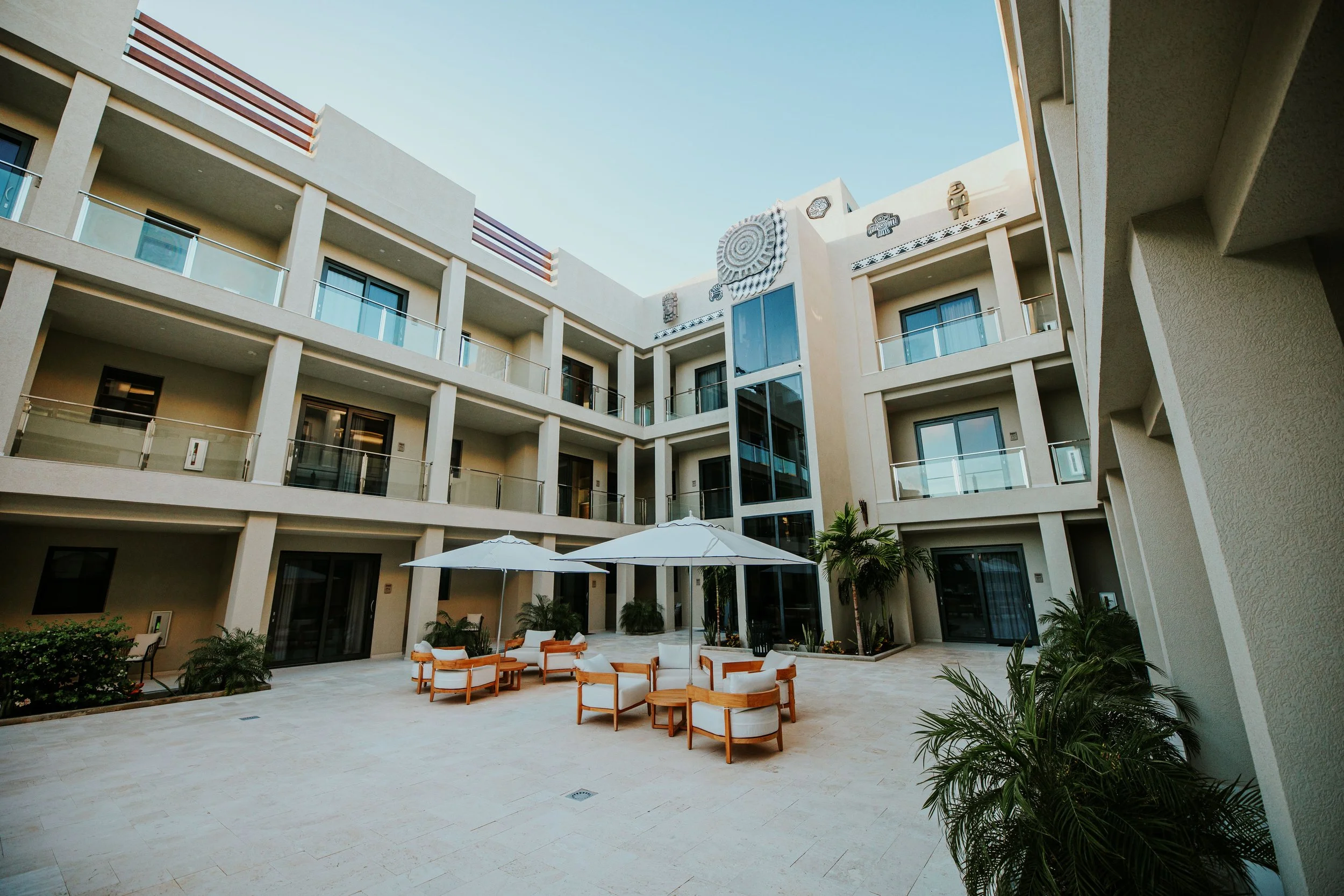 Courtyard of a modern hotel with outdoor seating, umbrellas, potted plants, and a multi-story building with balconies and decorative art on the top facade.