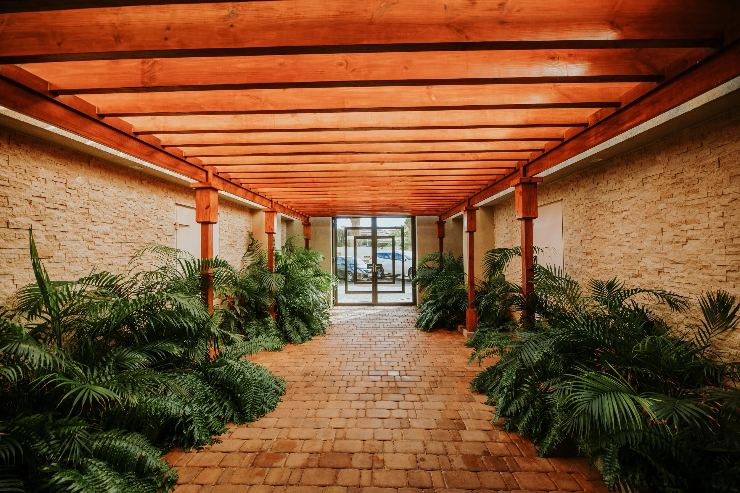 Indoor walkway with brick flooring, brick walls, wooden ceiling beams, potted green plants on both sides, and glass double doors at the end.