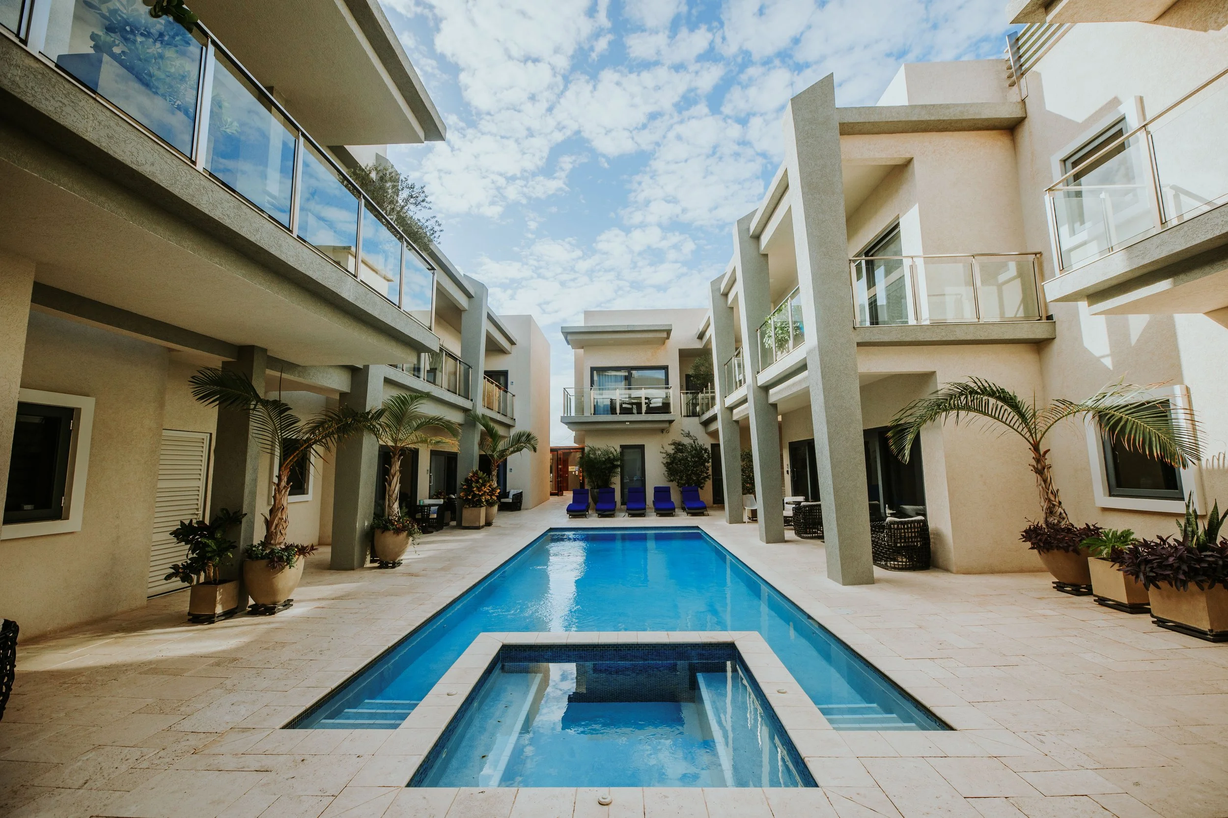 Courtyard of a modern apartment complex with a swimming pool, surrounded by beige buildings with balconies, potted plants, and blue lounge chairs, under a partly cloudy sky.