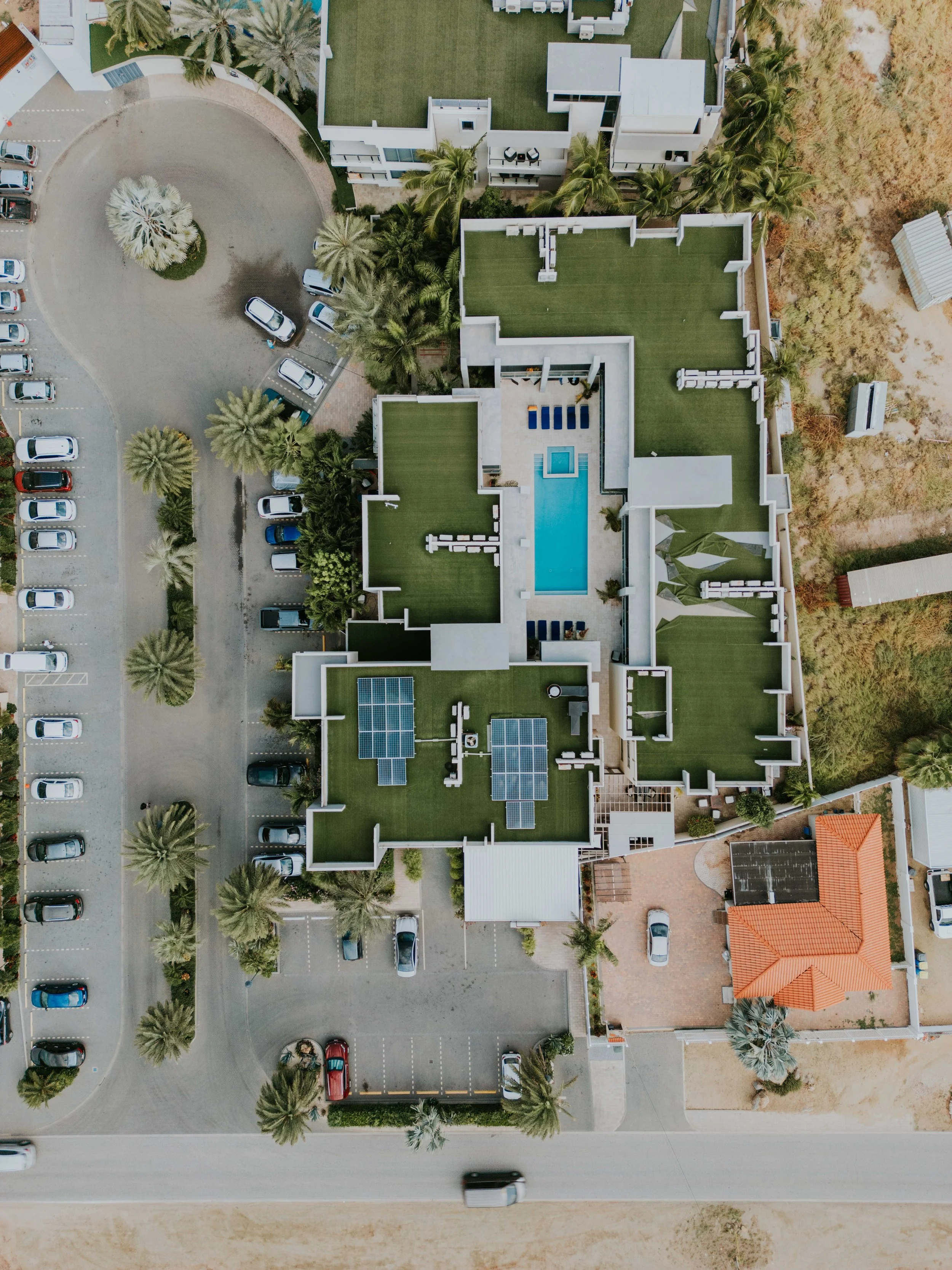 Aerial view of a modern apartment complex with a swimming pool, rooftop gardens with grass, solar panels, parking lot with cars, palm trees, and surrounding desert landscape.