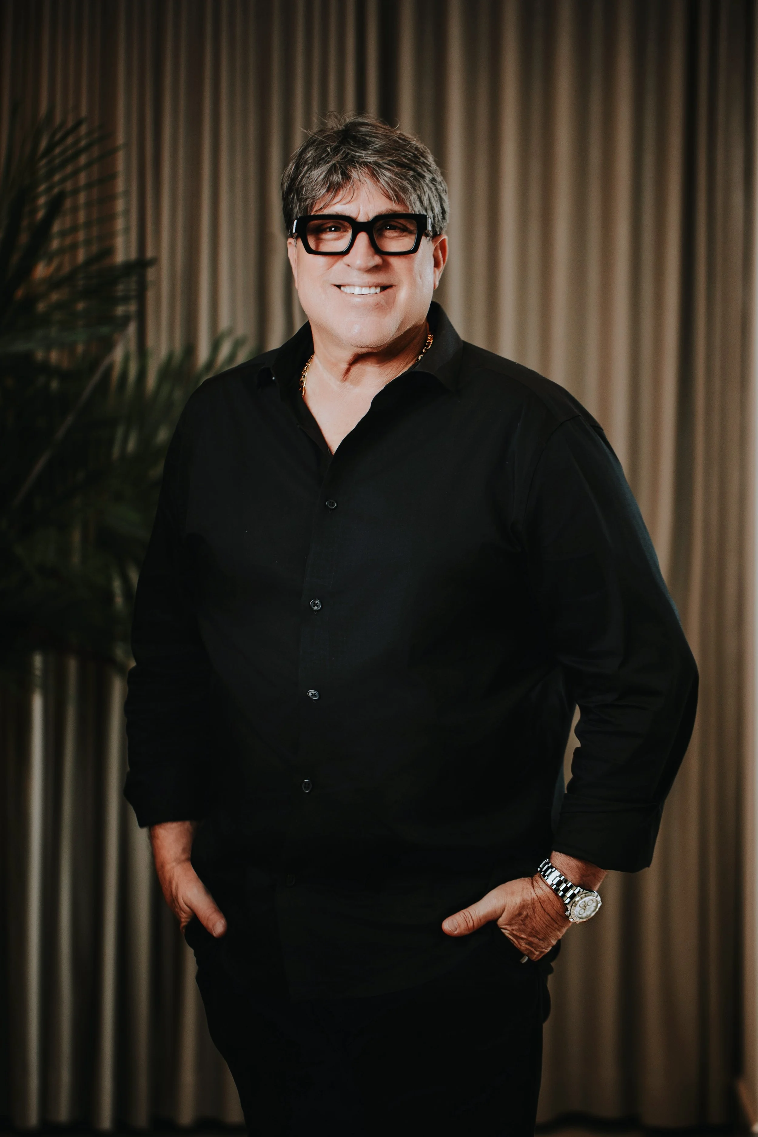 A man wearing a black shirt, glasses, and a watch, standing with one hand in his pocket in front of beige curtains.
