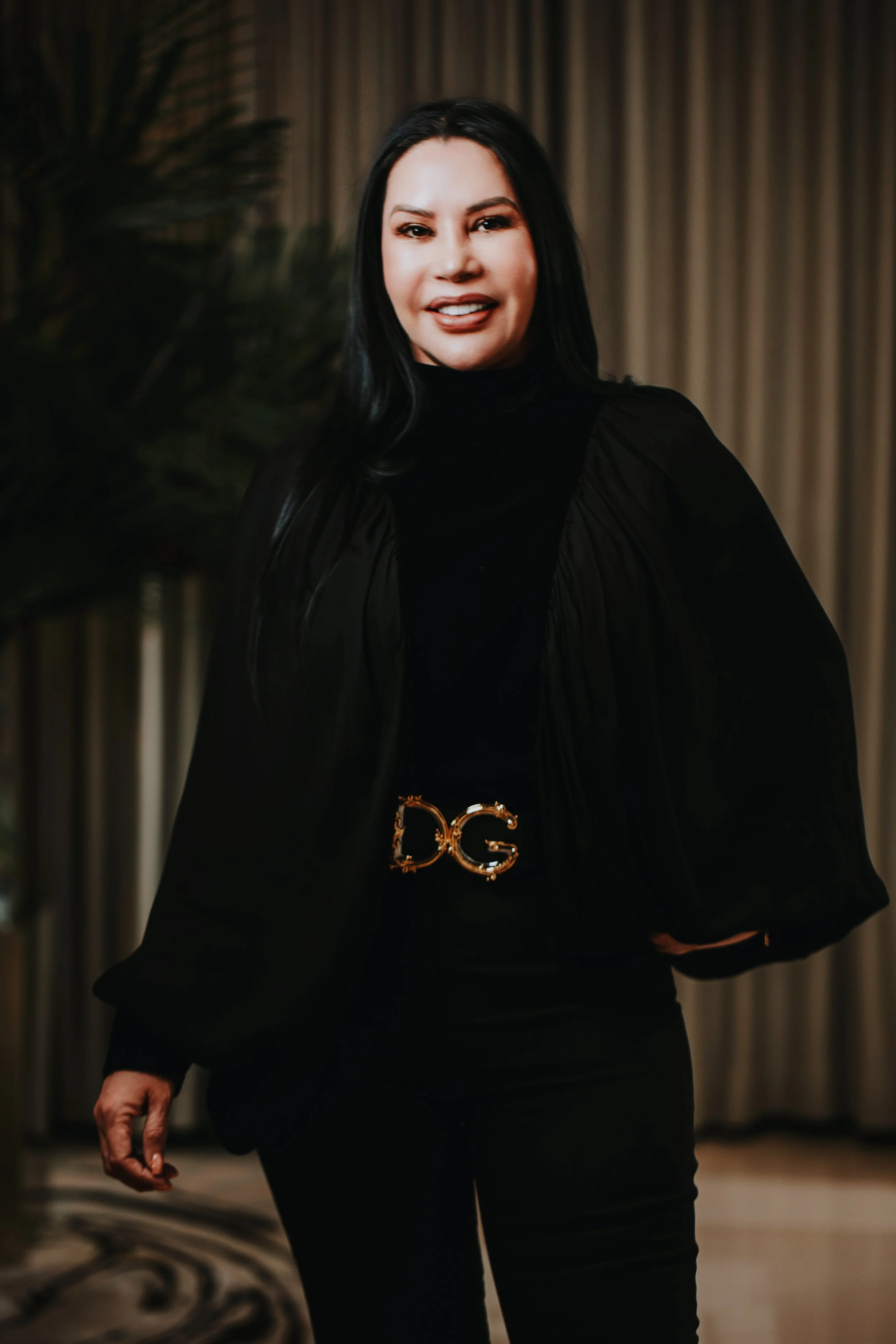 A woman with long black hair, wearing a black turtleneck and black pants, standing indoors with a curtain and plant in the background.